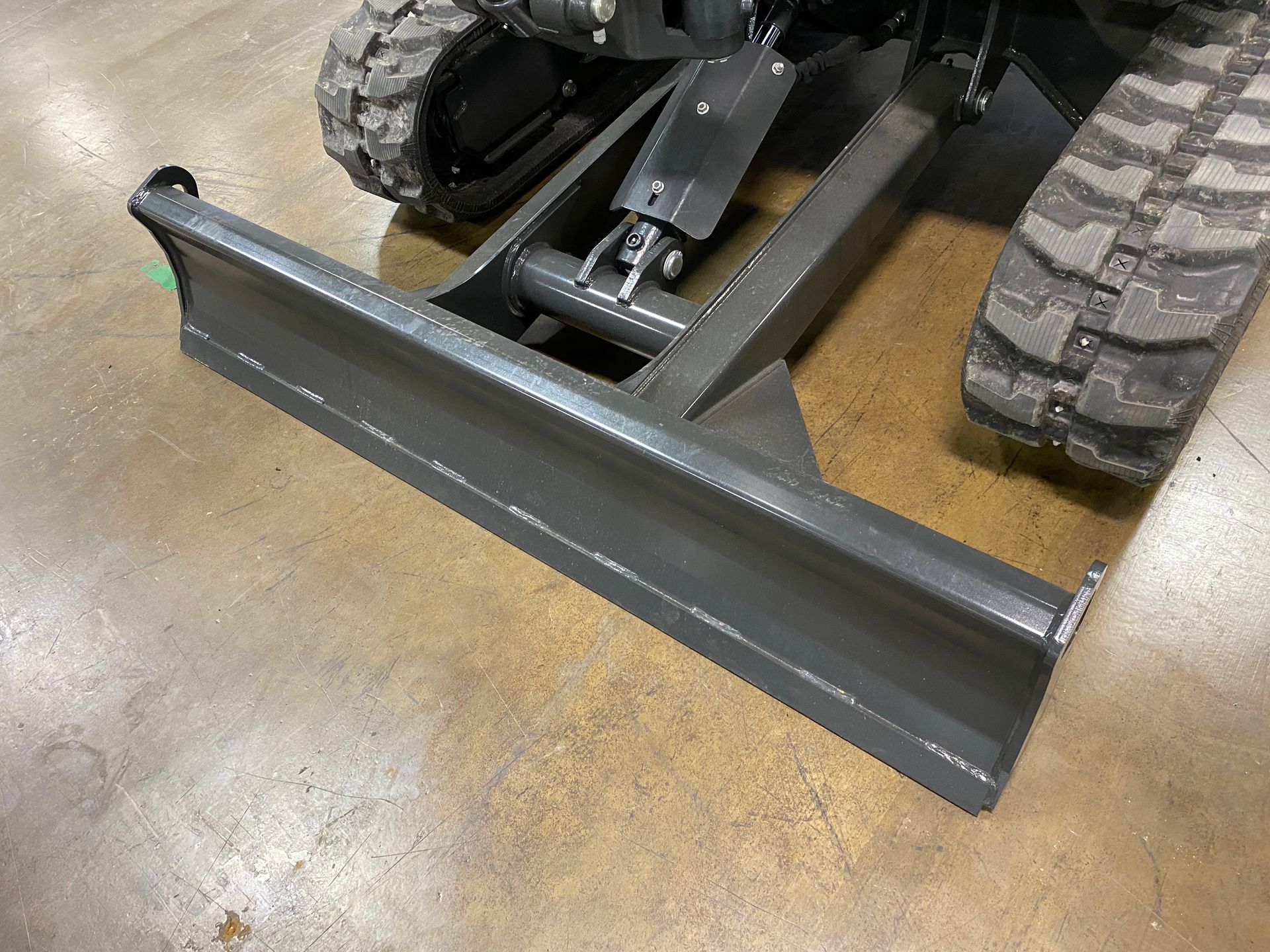 Gray bulldozer blade attached to a compact excavator, on a concrete floor.