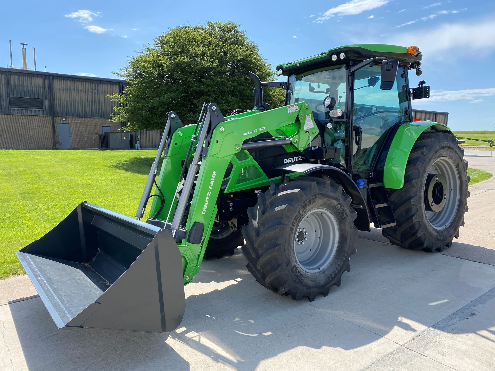 Green tractor with front loader on concrete, parked near a building and tree under a blue sky.