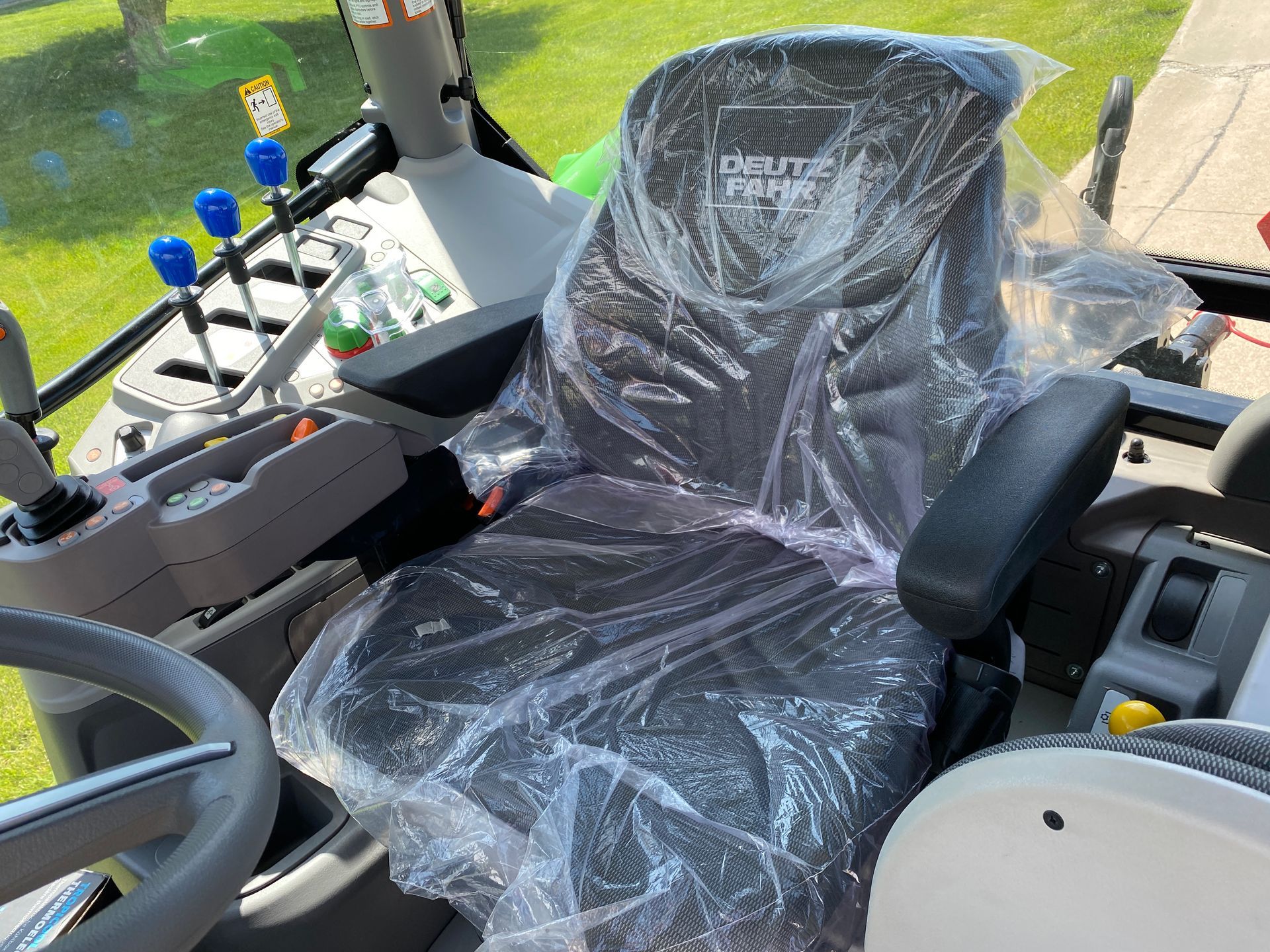 Interior of a Deutz-Fahr tractor, driver's seat covered in plastic. Steering wheel, controls, and levers visible.