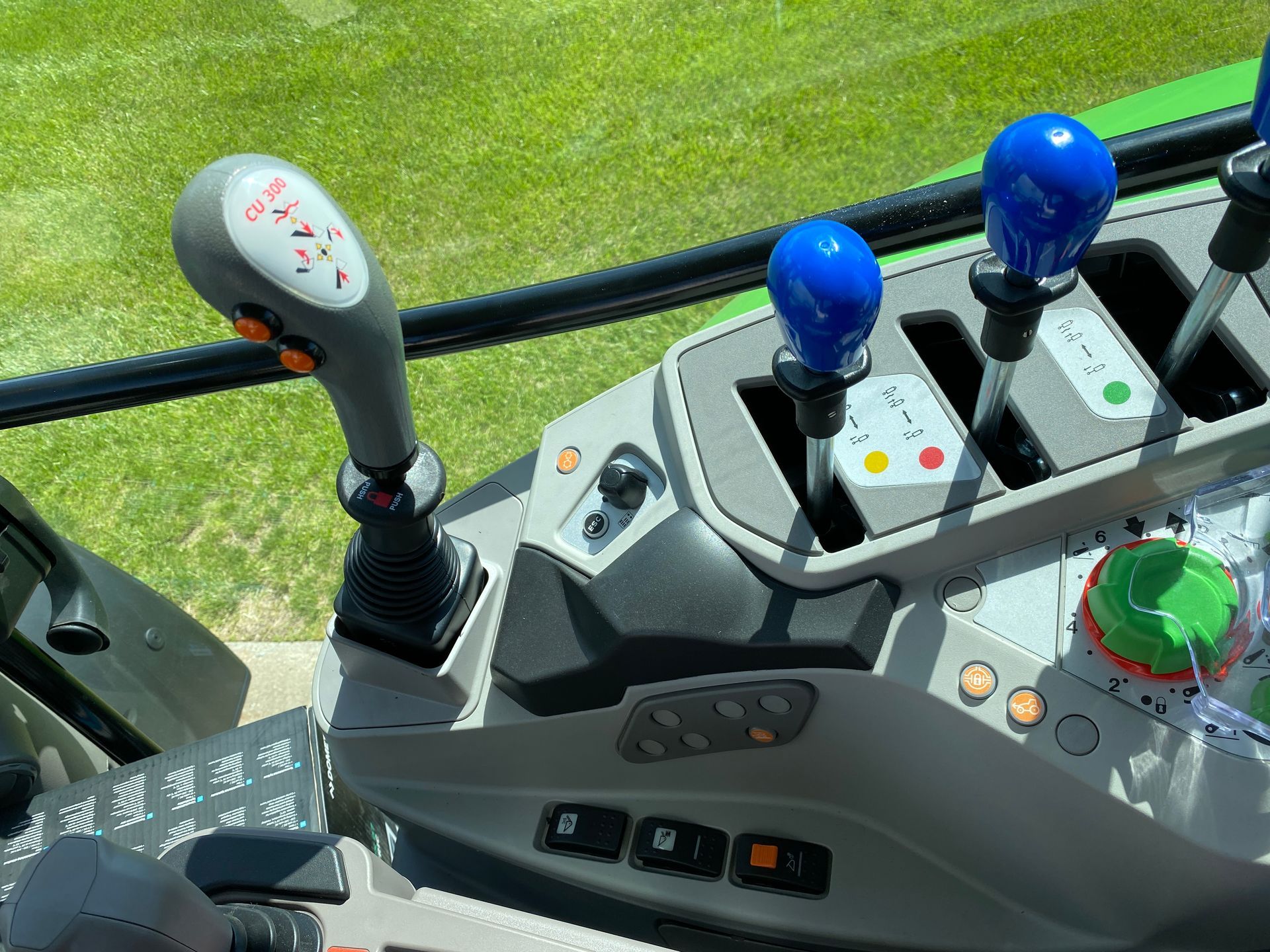 Close-up of a tractor cab's control panel, showing multiple levers and buttons, with a green field visible in the background.
