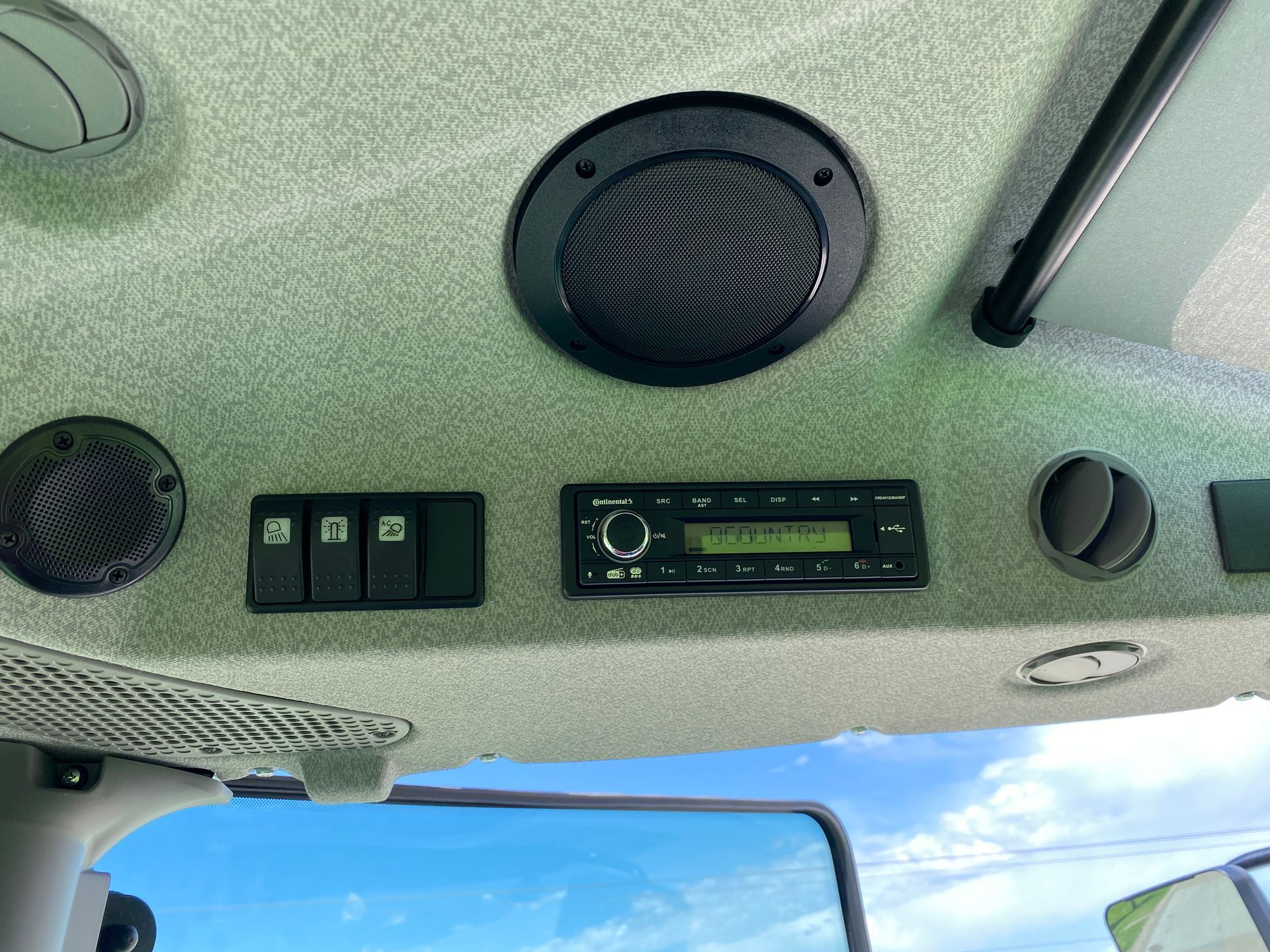 Interior of a vehicle's ceiling with speakers, radio, and switches. Green and black with a blue sky visible.