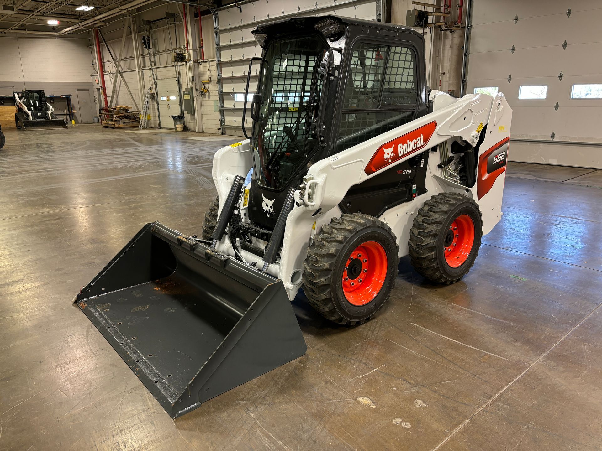 White Bobcat skid-steer loader with black cab, gray bucket, and orange wheels sits on a concrete floor in a warehouse.