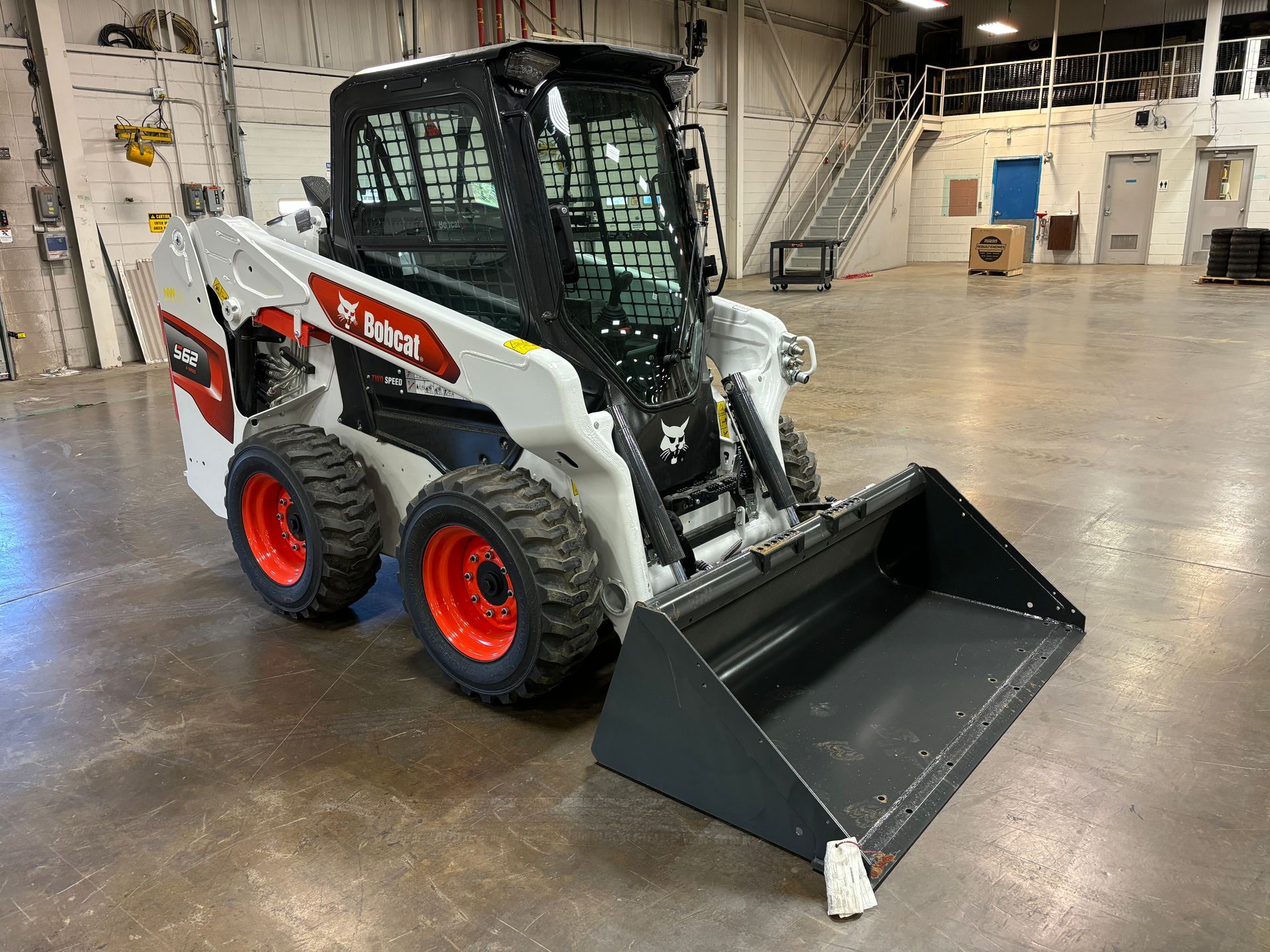 White and black Bobcat skid-steer loader with a gray bucket, parked inside a warehouse.