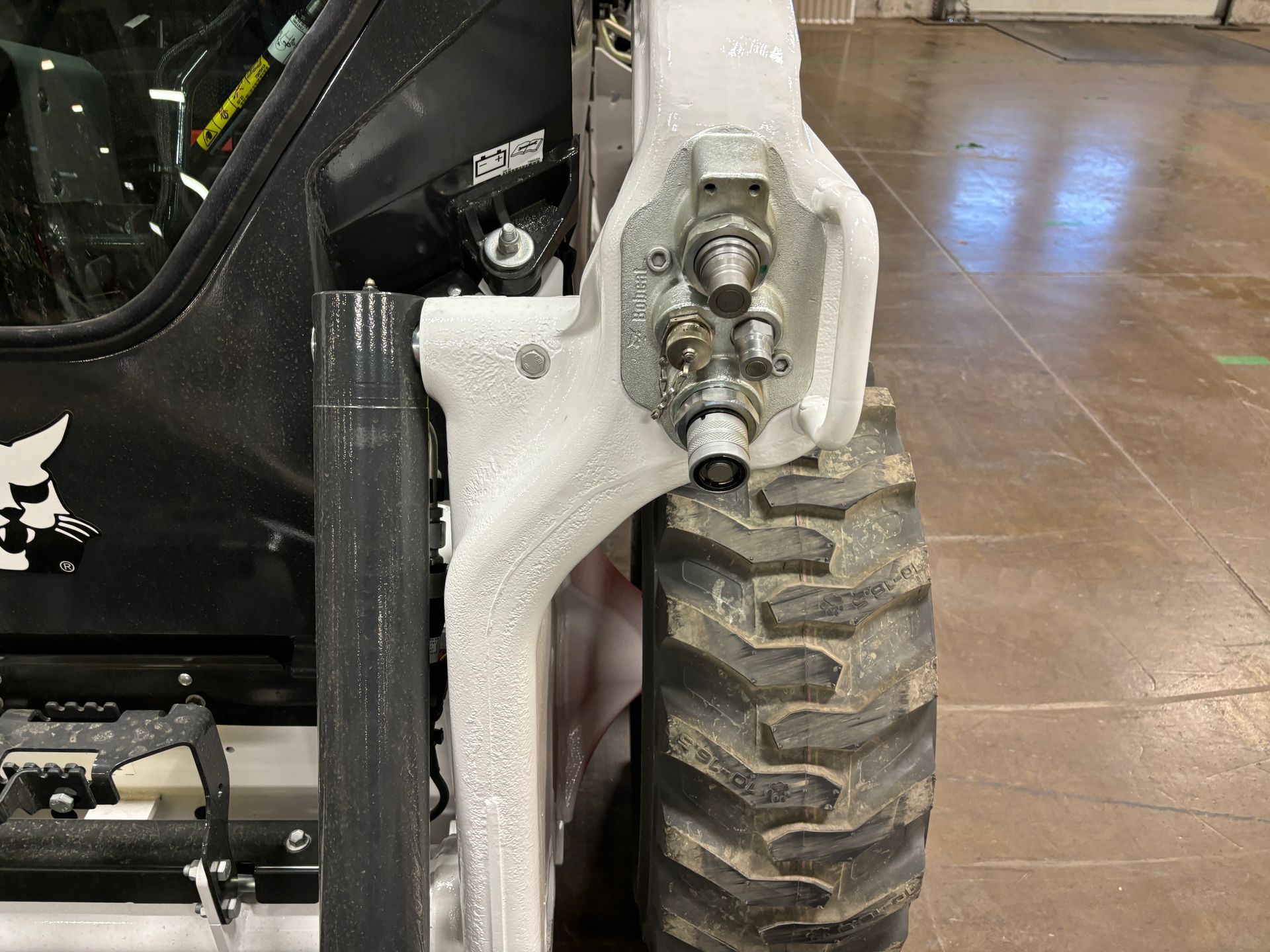 A close-up of a Bobcat skid steer’s wheel, hydraulic fittings, and cab, showing a snowy environment.