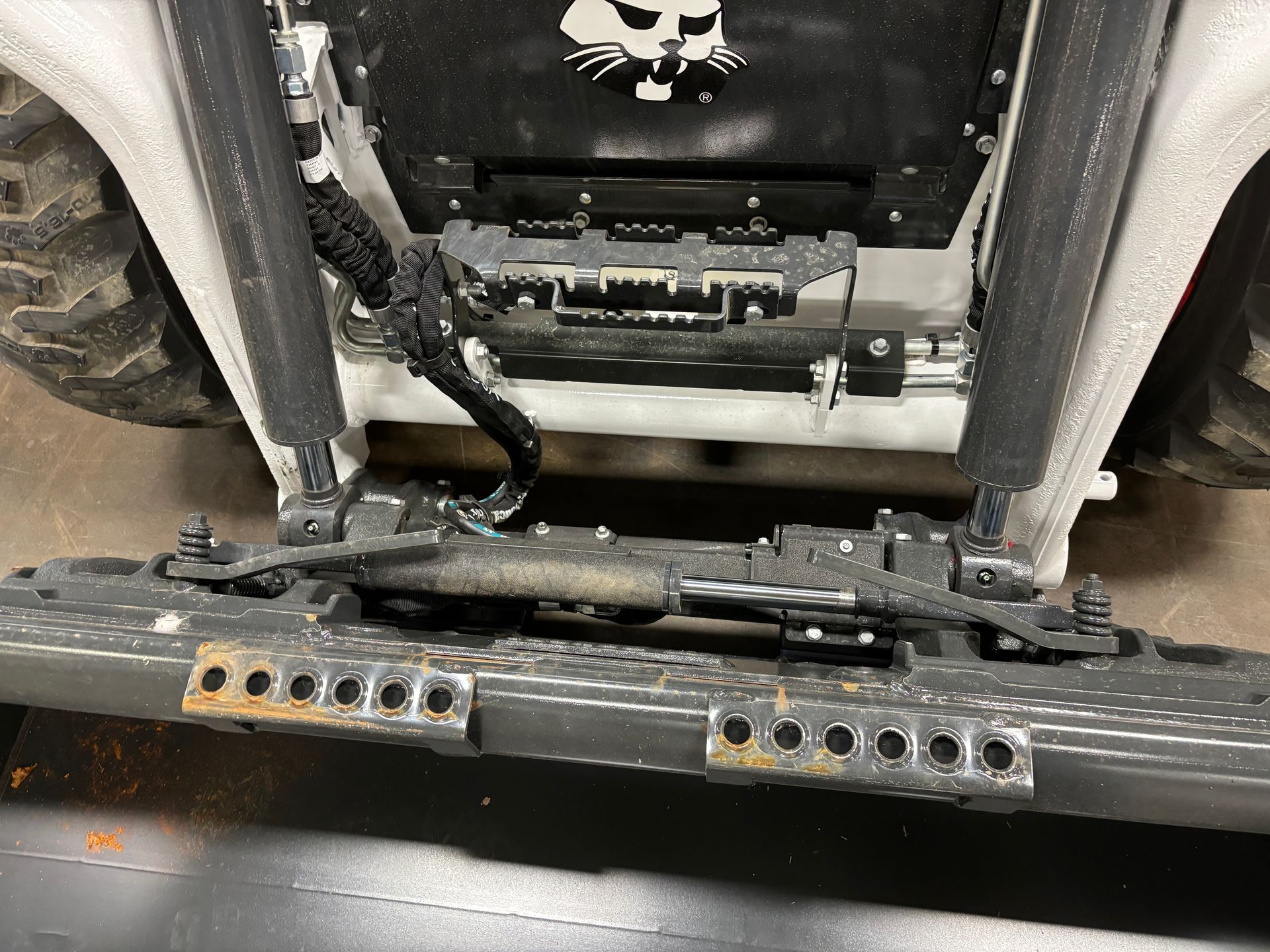 Underside of a vehicle showing rusty metal step, suspension parts, and wiring. A black panel with a white cat skull logo is visible.