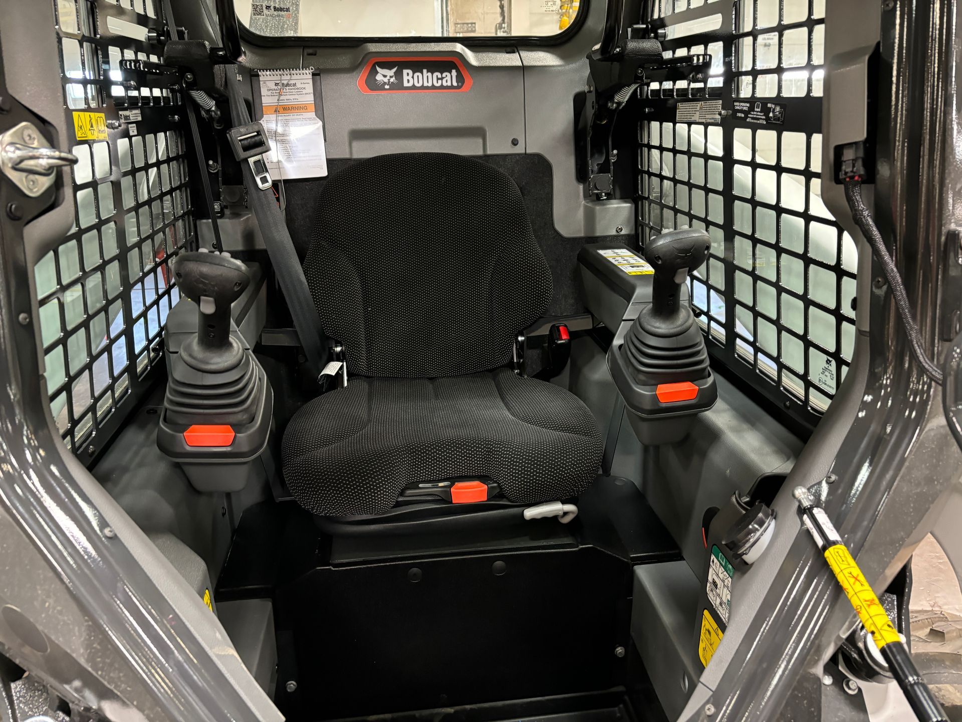 Interior view of a Bobcat skid-steer loader cab, with seat, joysticks, safety cage, and control panel.