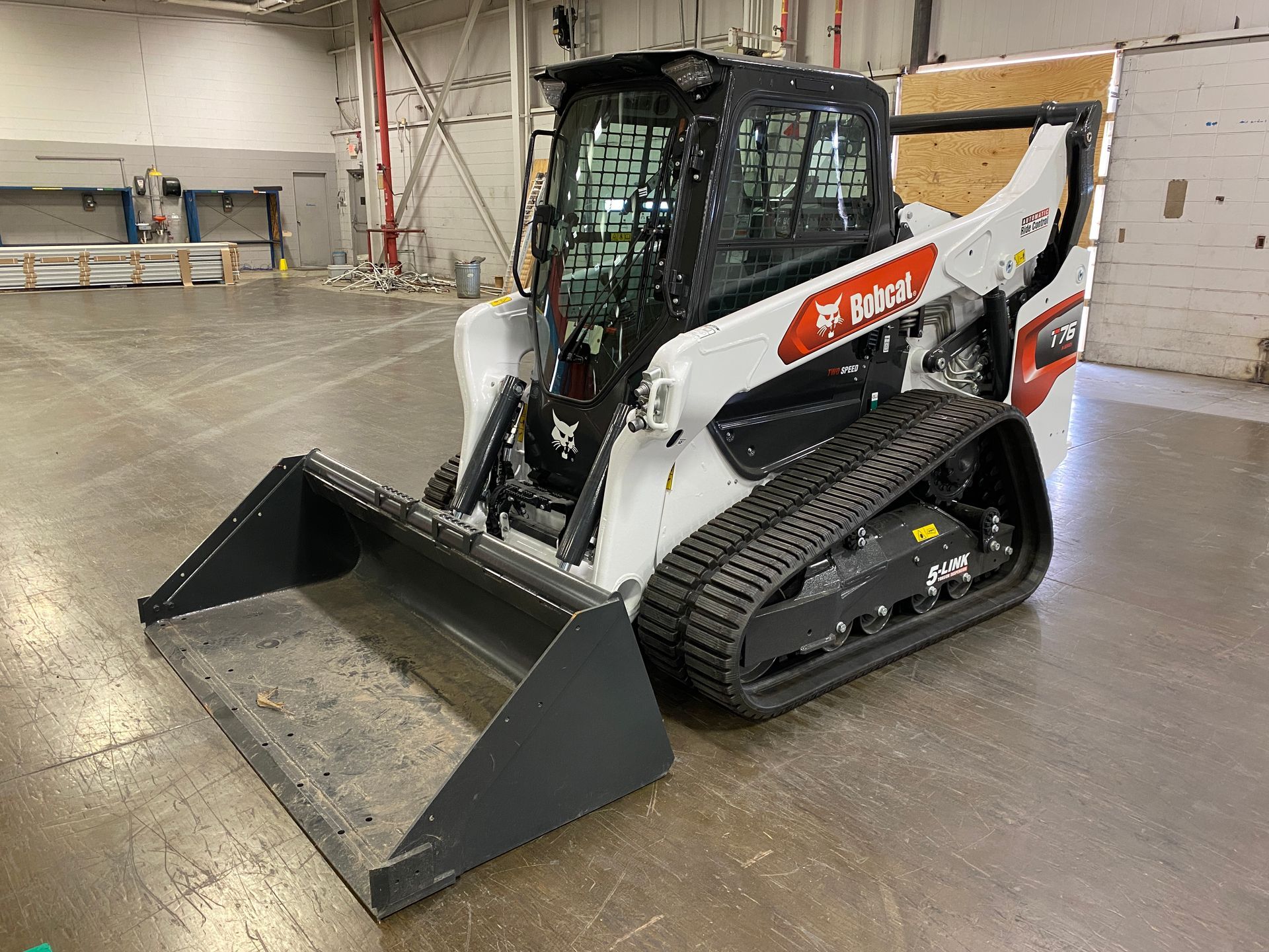 White and red Bobcat track skid-steer loader with a front bucket parked on a smooth floor inside a building.