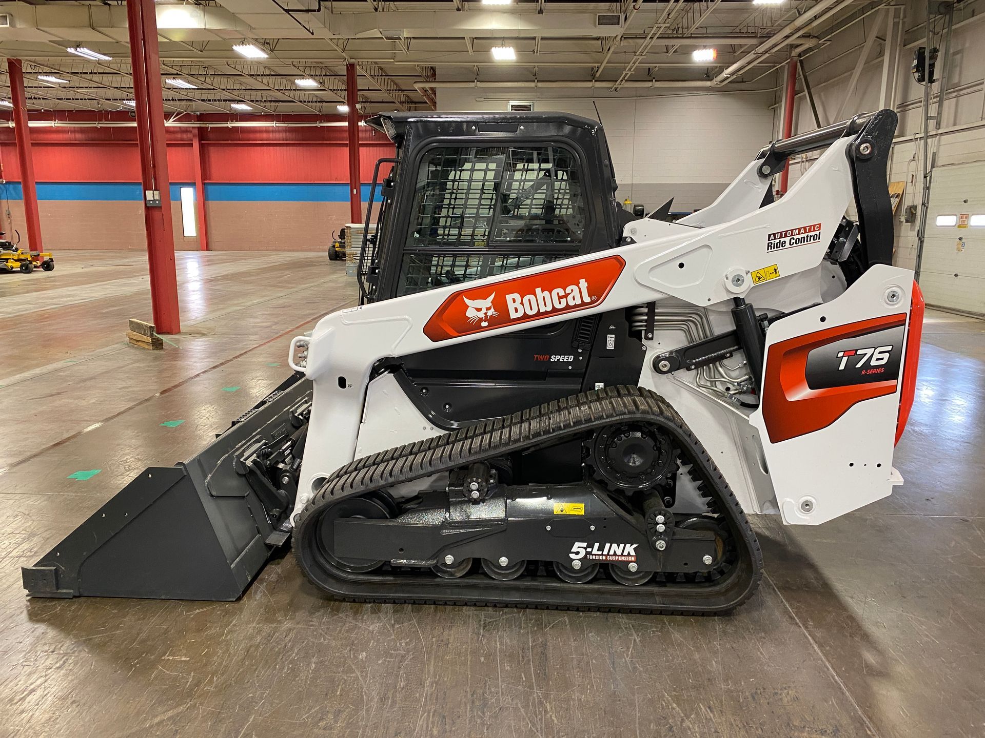White and orange Bobcat T76 compact track loader with a bucket in an indoor warehouse.