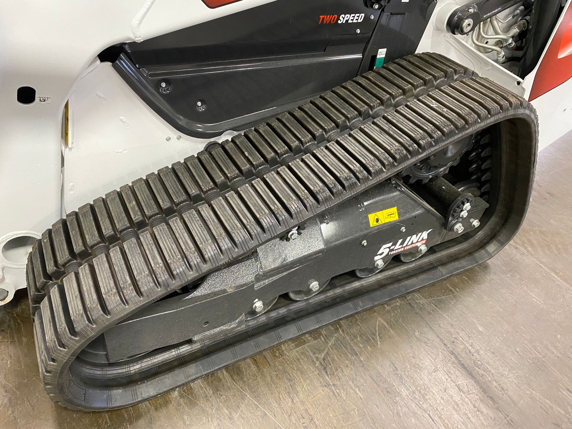 Track of a white and gray skid steer loader on a wood floor, showing the tread.