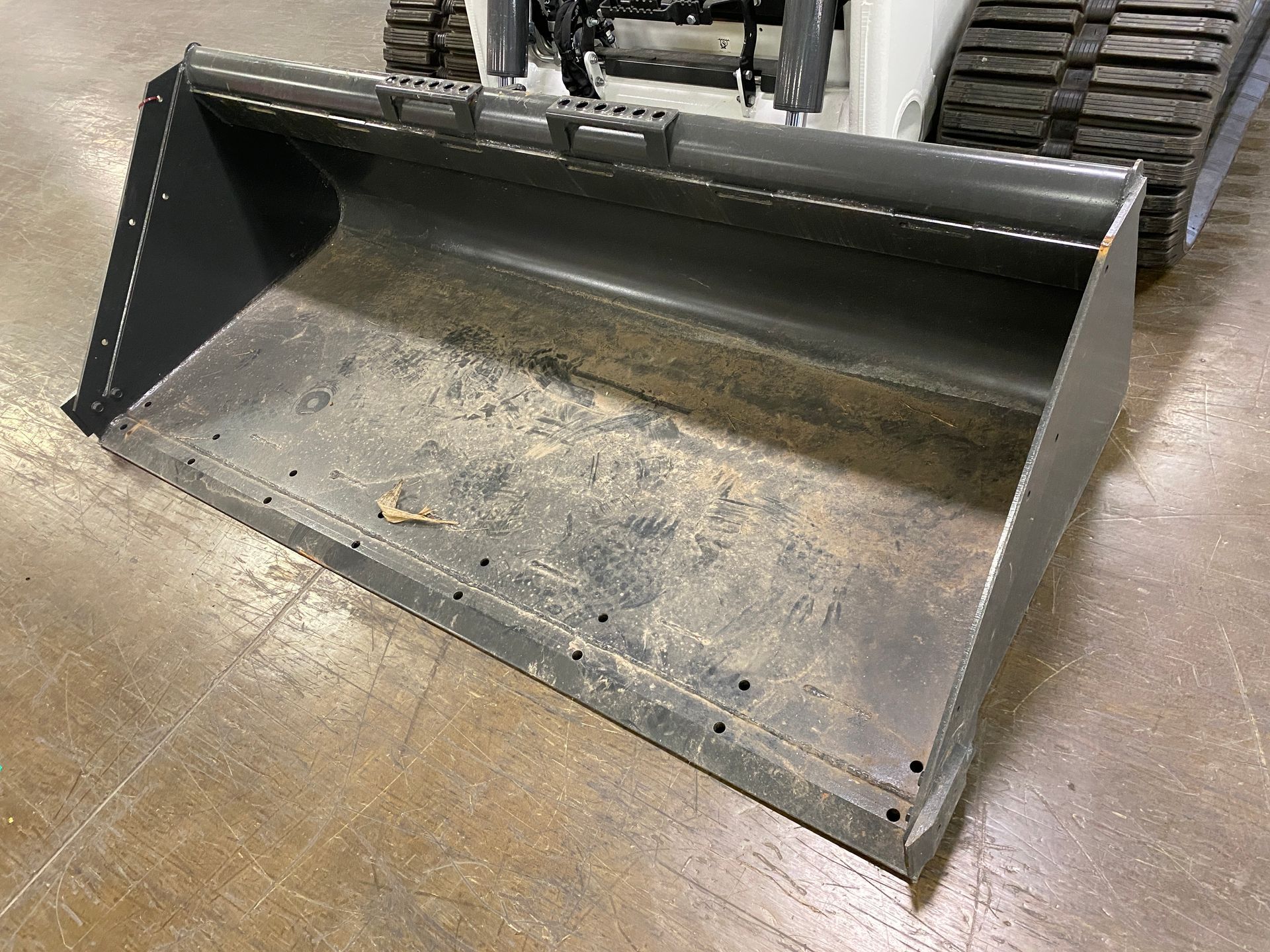 Gray metal bucket attached to a small white construction vehicle, sitting on a wooden floor.