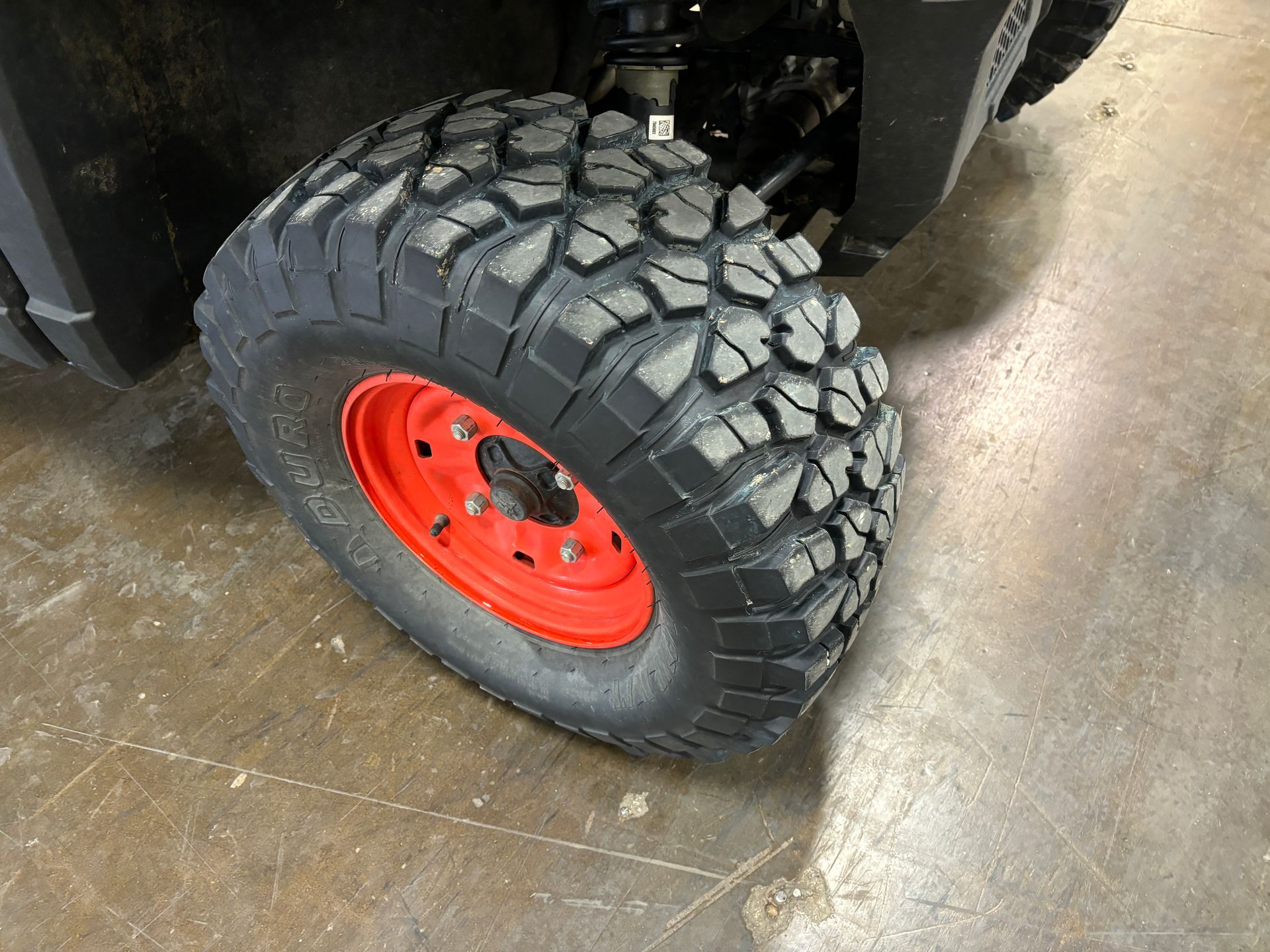 Black off-road tire on a red rim, attached to a vehicle. The setting appears to be a concrete floor.