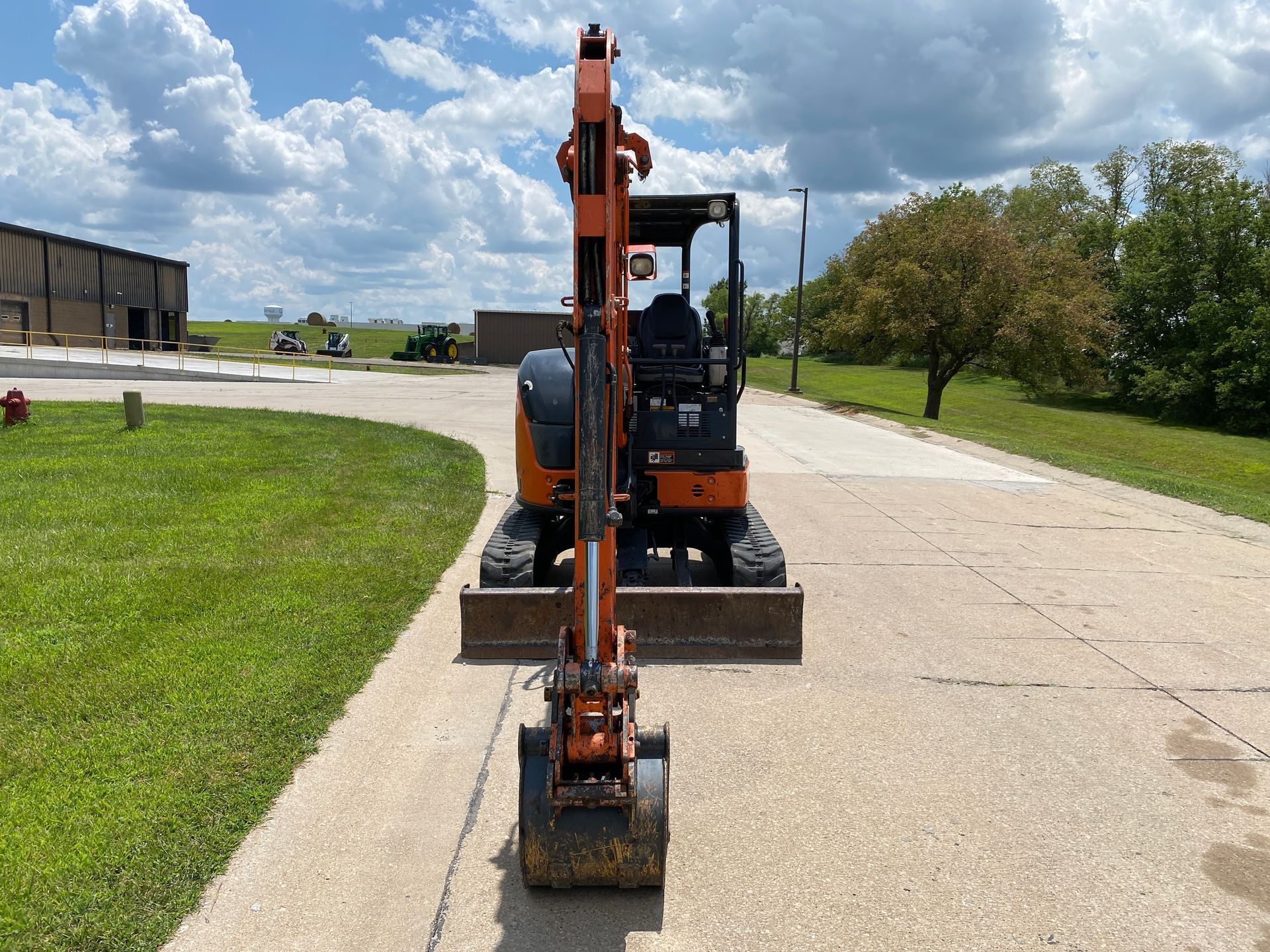 Orange mini-excavator on pavement, facing forward. Grass and a building are in the background under a blue sky.