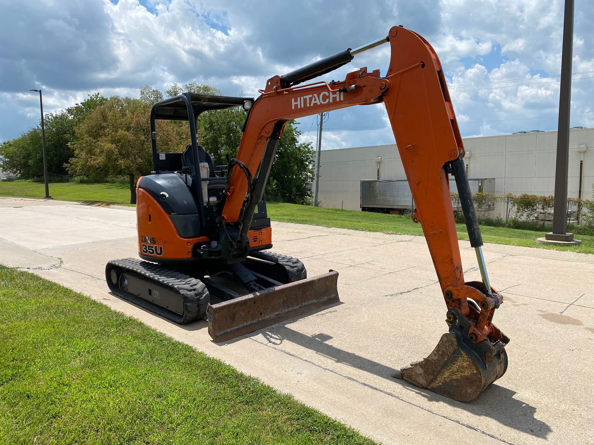 Orange Hitachi mini excavator on pavement beside grass, cloudy sky background.