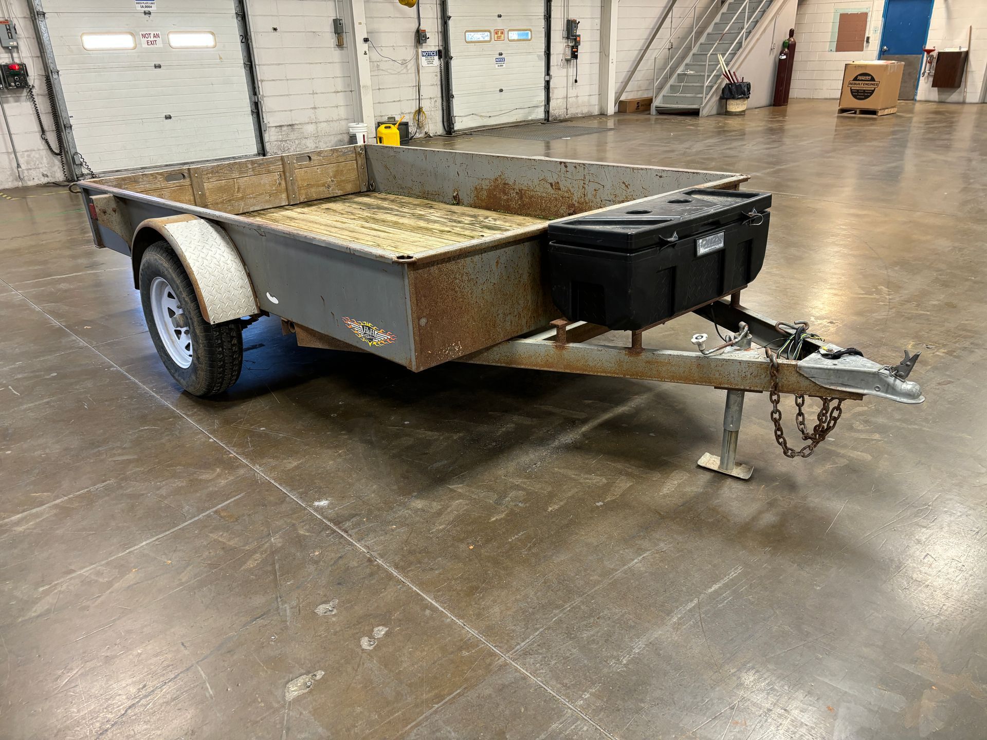 Grey utility trailer with a black storage box attached, in a large warehouse.