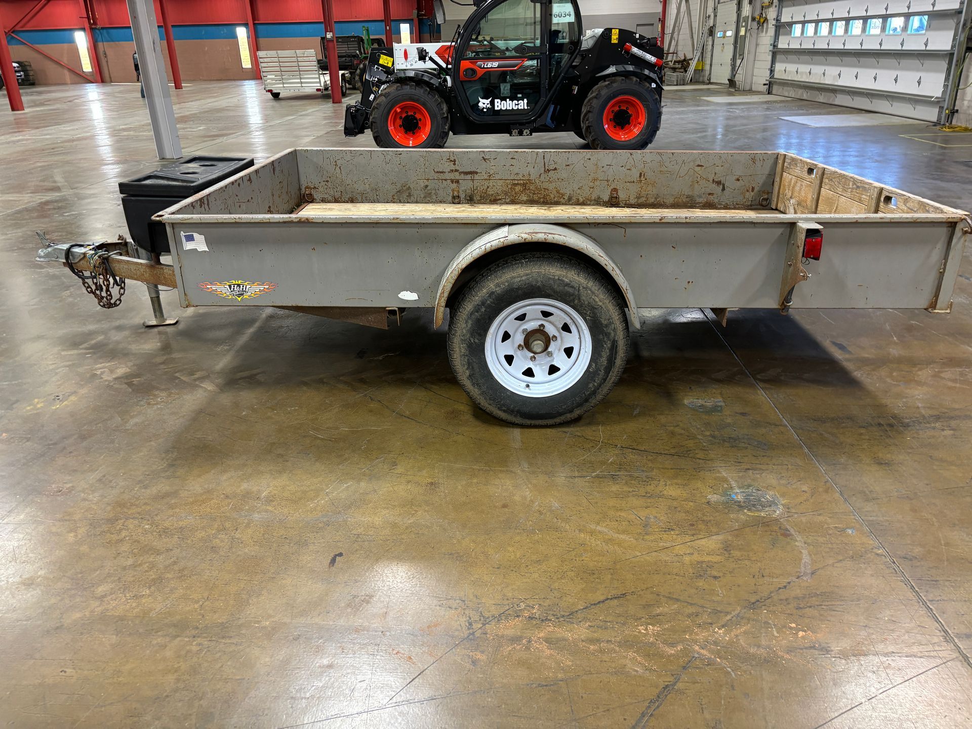 A gray utility trailer parked on concrete with a Bobcat loader in the background.