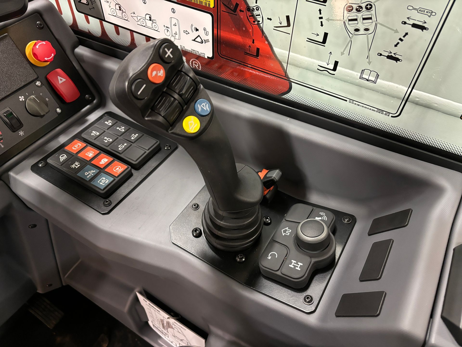 Control panel with joystick, buttons, and switches in a construction vehicle. Gray and black with red emergency stop button.