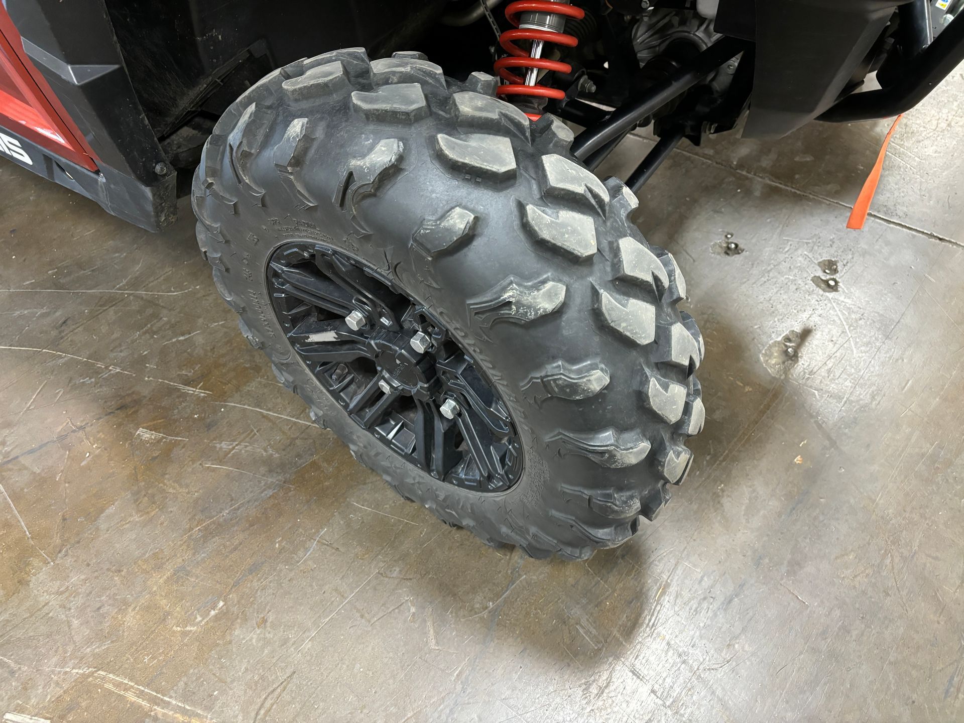 Off-road vehicle tire with chunky tread, black wheel, red suspension, angled on a concrete surface.