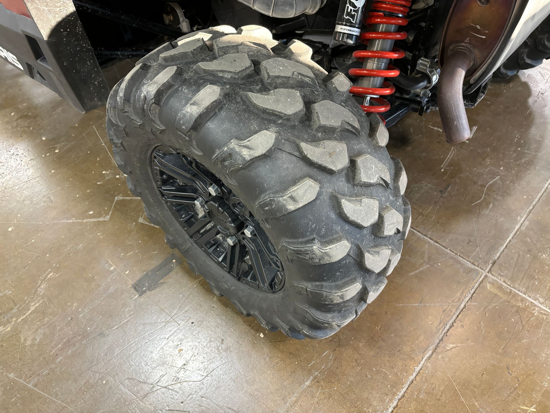Off-road vehicle tire with deep tread, on concrete floor. Red suspension visible.
