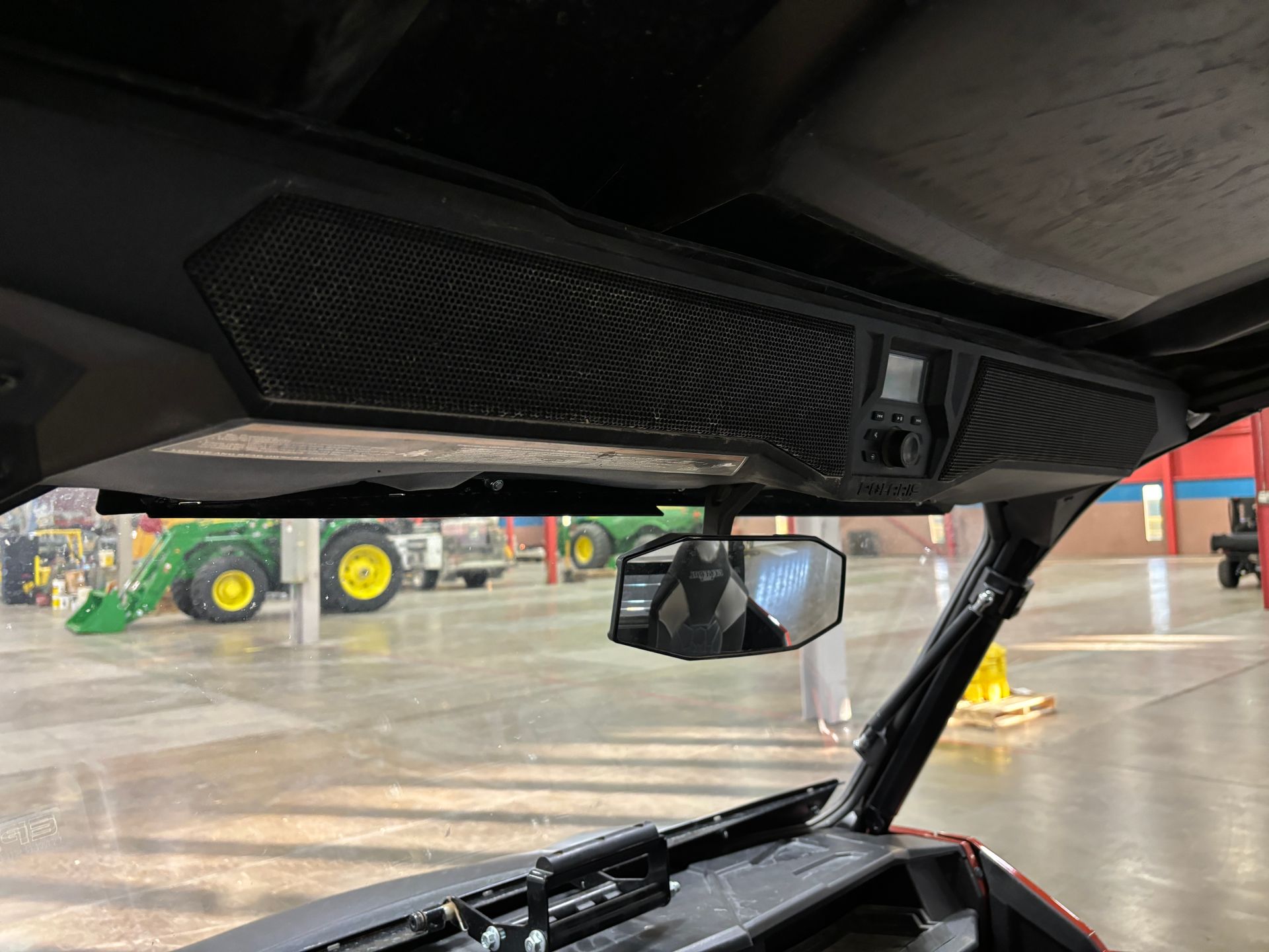 Interior view of a side-by-side vehicle with a mesh speaker console mounted to the roof. A rearview mirror hangs below.