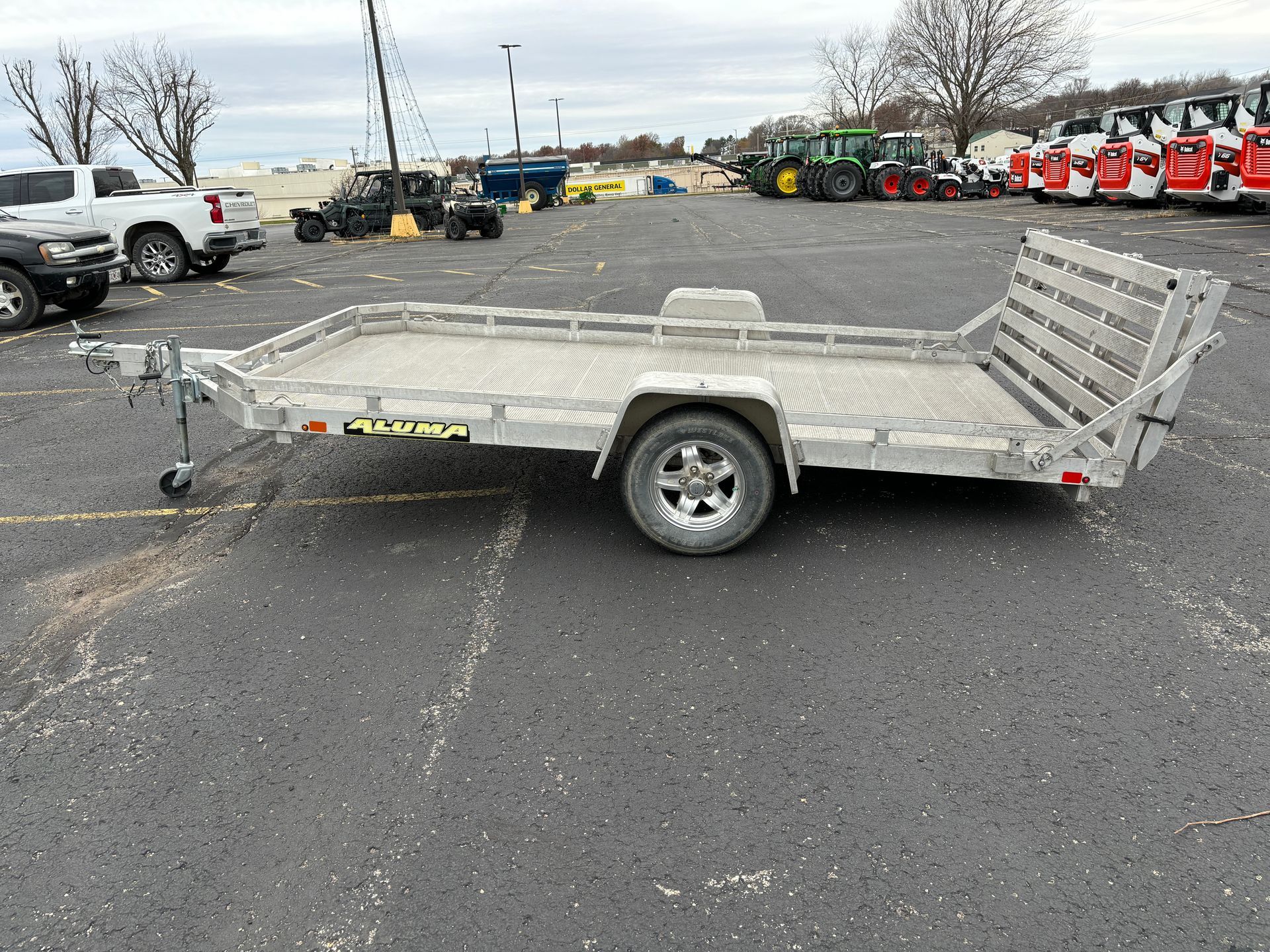 Aluminum utility trailer on asphalt parking lot, vehicles and buildings in background.