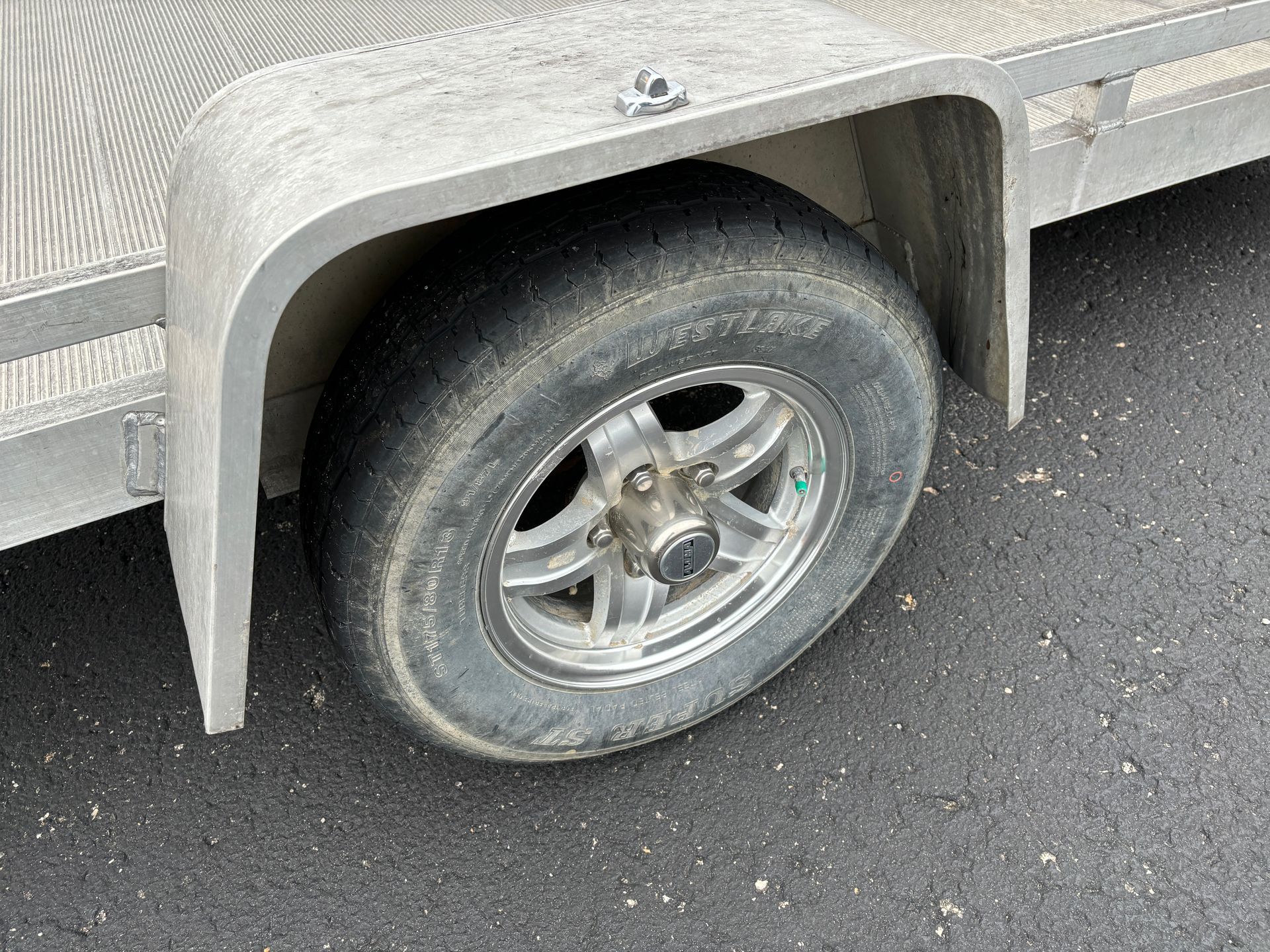 Trailer wheel with a gray tire and silver rim, set against an asphalt surface.