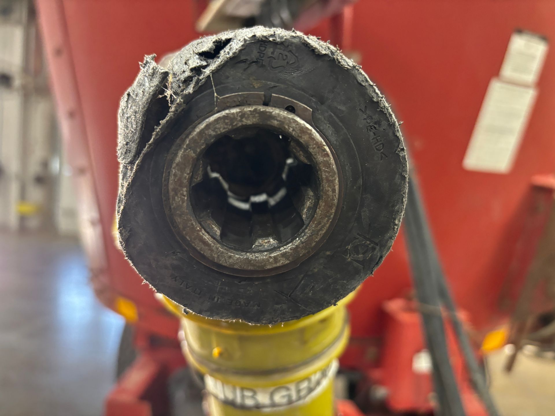 Close-up of a damaged agricultural shaft with a chewed-up black rubber cover. Yellow and red machinery are visible.
