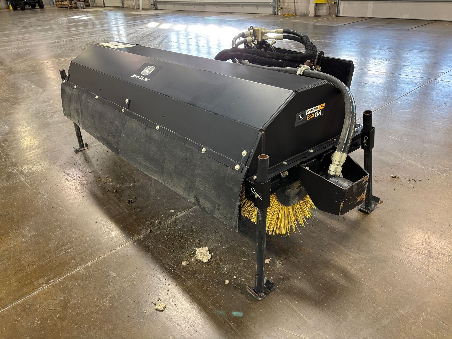 Black John Deere angle broom with yellow bristles on a concrete floor at an angle.