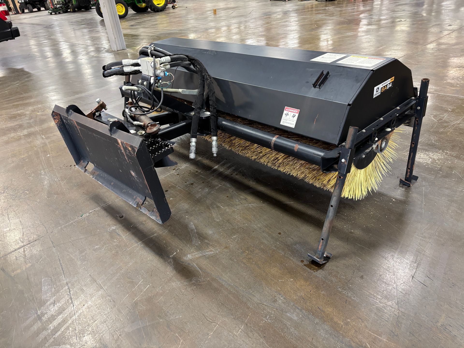 Black John Deere angle broom with yellow bristles on a concrete floor.