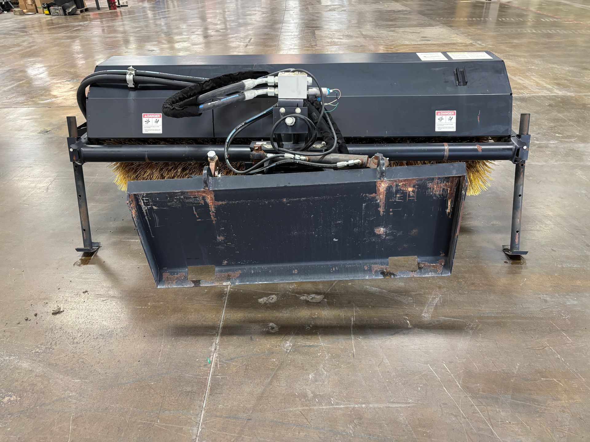 Black John Deere angle broom with yellow bristles on a concrete floor at a back view.