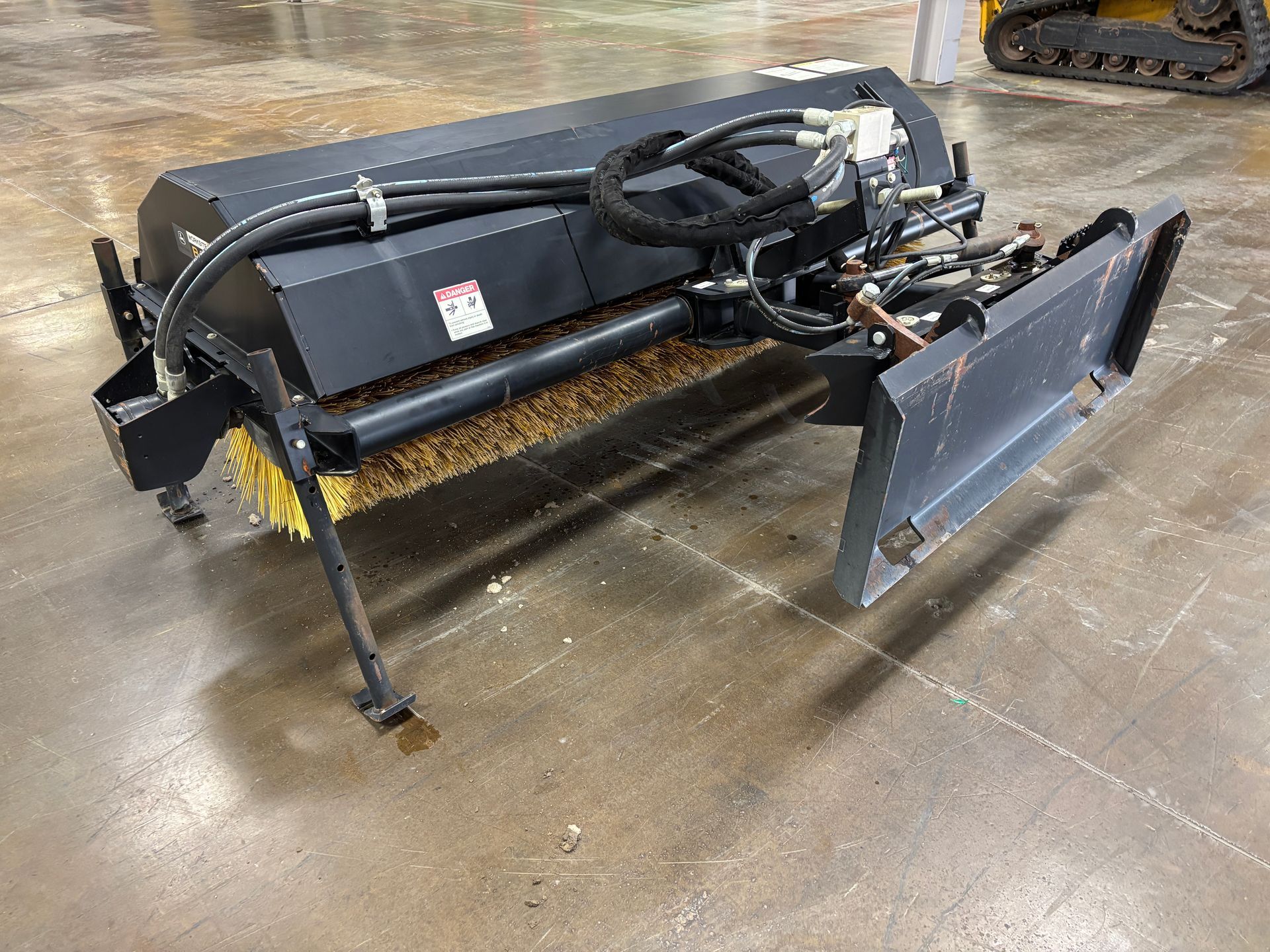 Black John Deere angle broom with yellow bristles on a concrete floor.