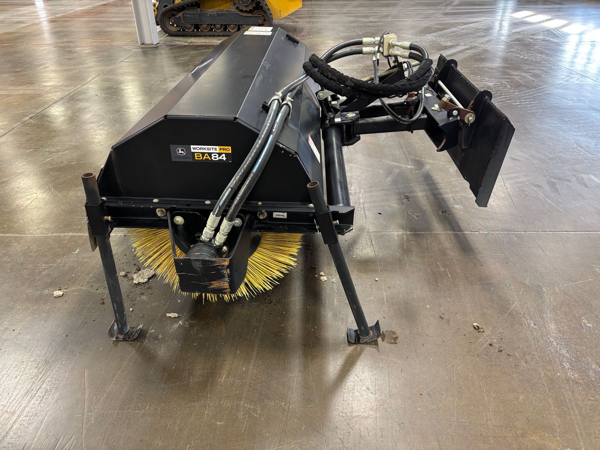 Black John Deere angle broom with yellow bristles on a concrete floor.