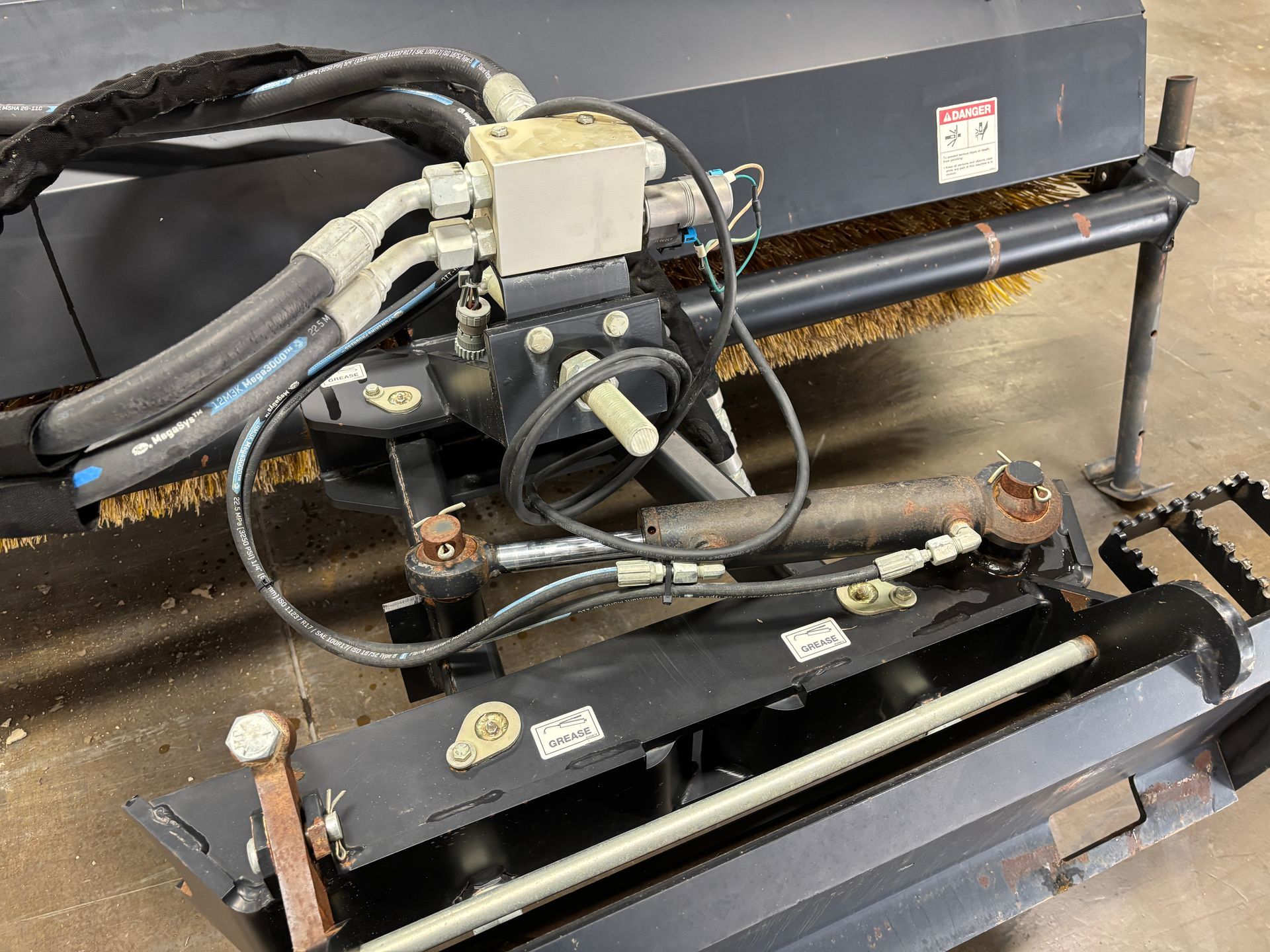 Black hydraulic-powered street sweeper brush mechanism. Hoses, valves, and a control box are visible.