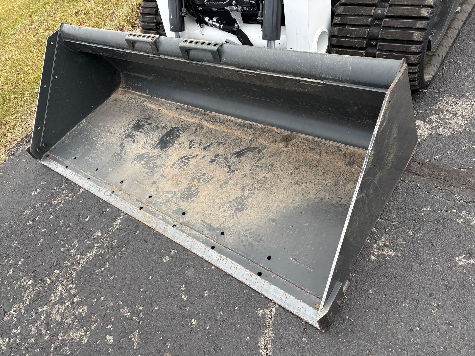 Black bucket of a compact track loader, outdoors on asphalt, filled with dirt.