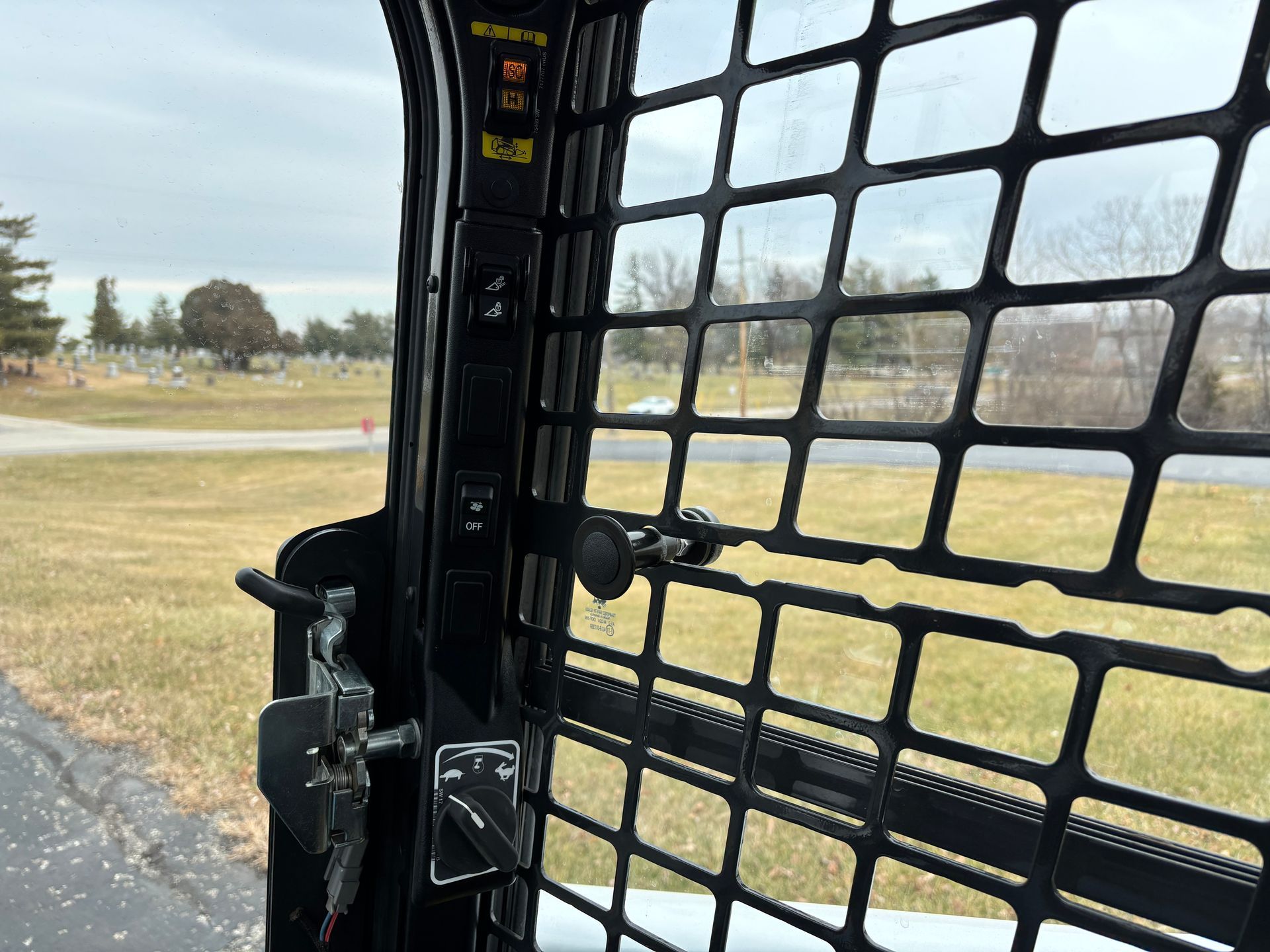 Interior of a compact track loader cab with black safety grill and controls. Exterior view of a road and trees.