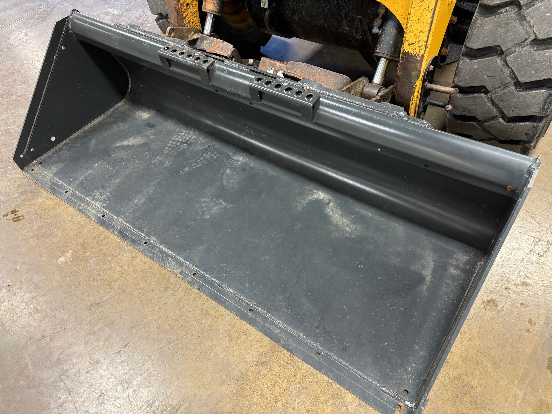 A gray front-end loader bucket attached to a yellow skid steer on a concrete surface.