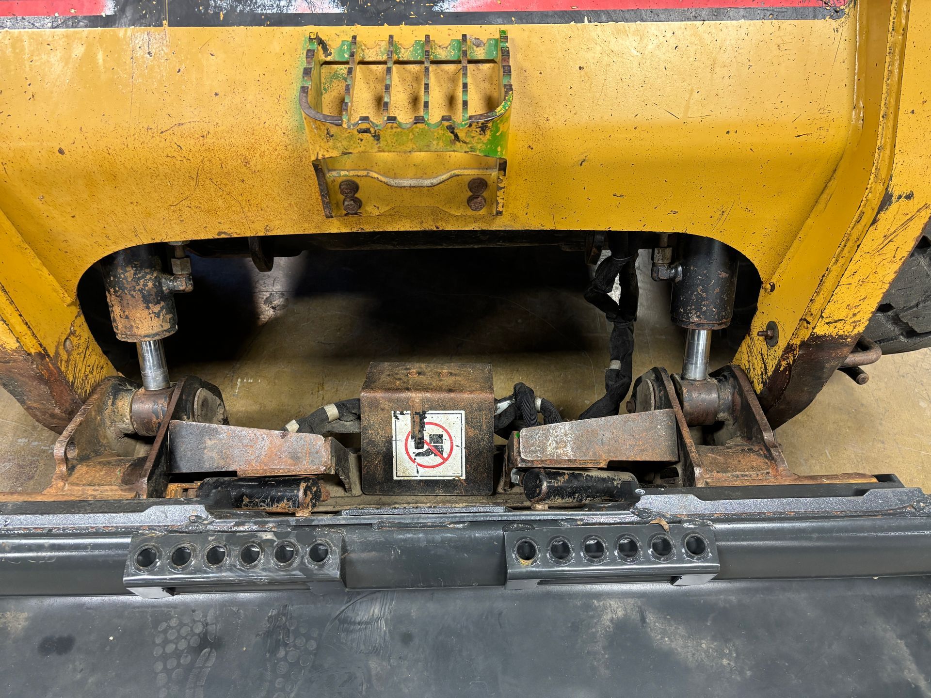 Rear view of a yellow skid steer loader with black bucket attachment. Visible hydraulic cylinders and safety warning.