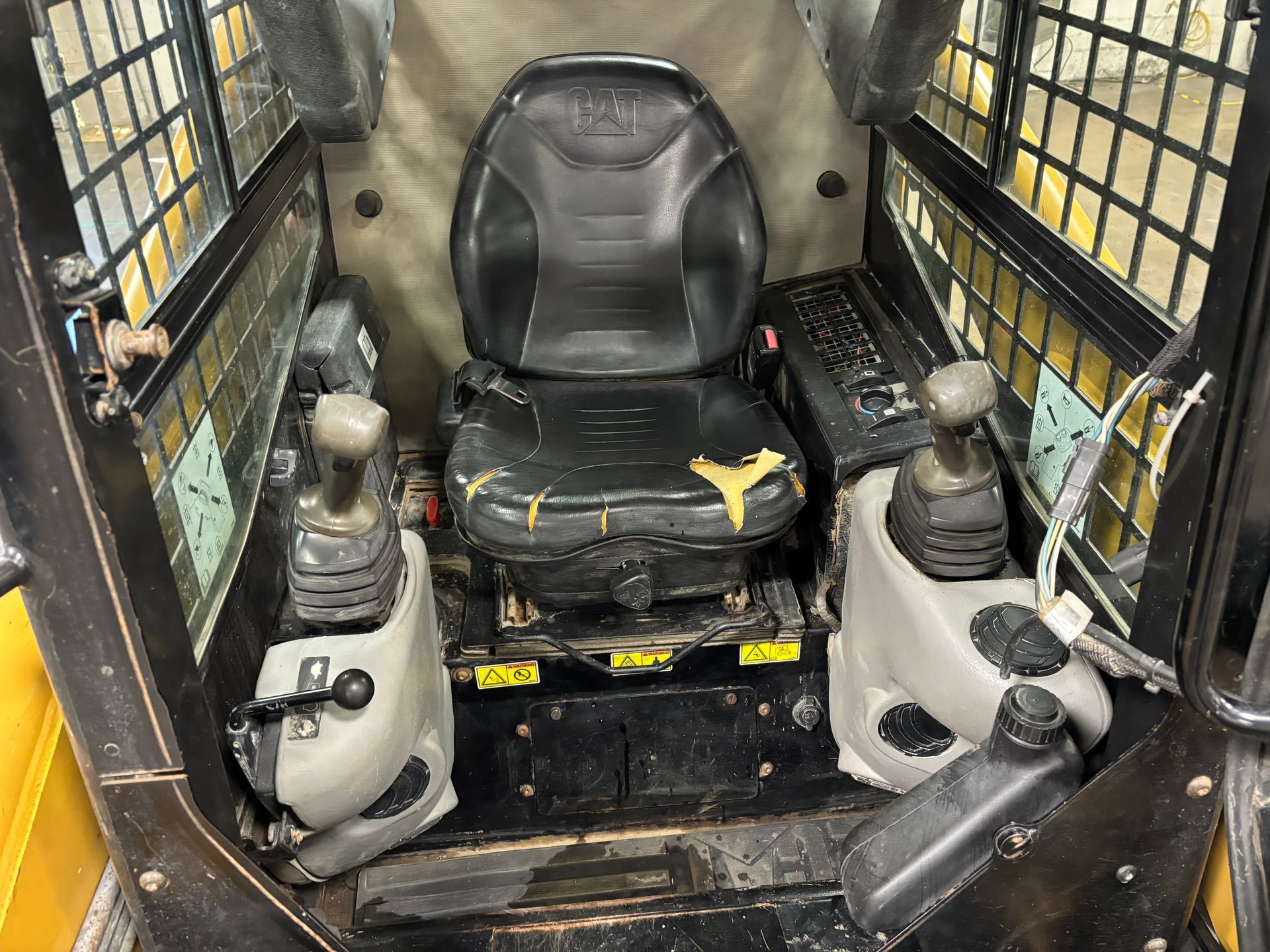 Interior of a skid steer loader cab, featuring a worn black seat, control levers, and safety bars.