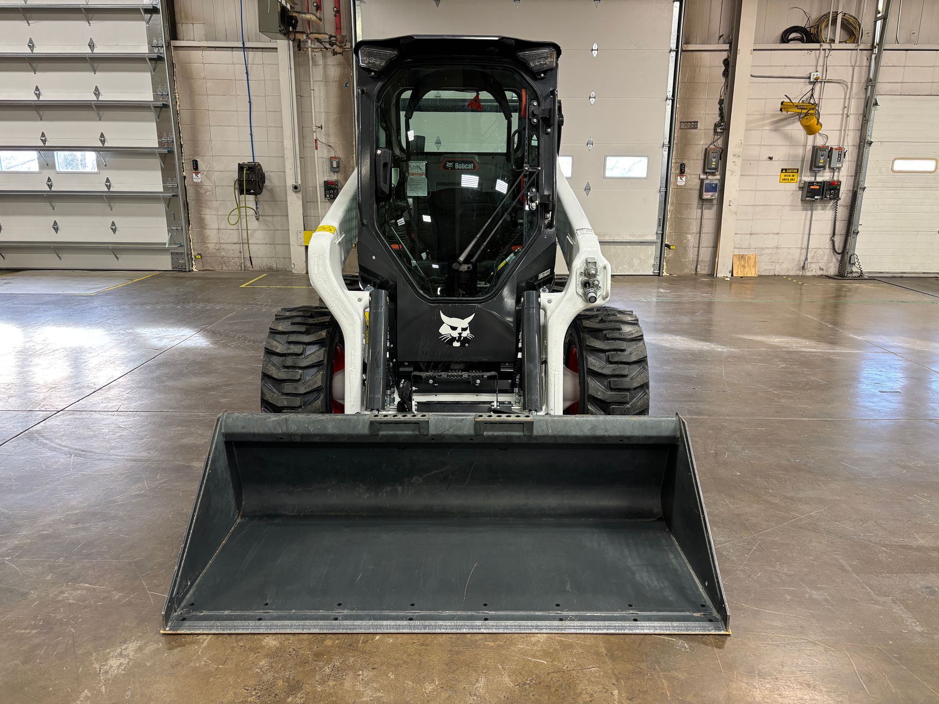 Bobcat skid-steer loader with a black bucket, indoors. White cab, black tires, parked in front of a closed garage door.