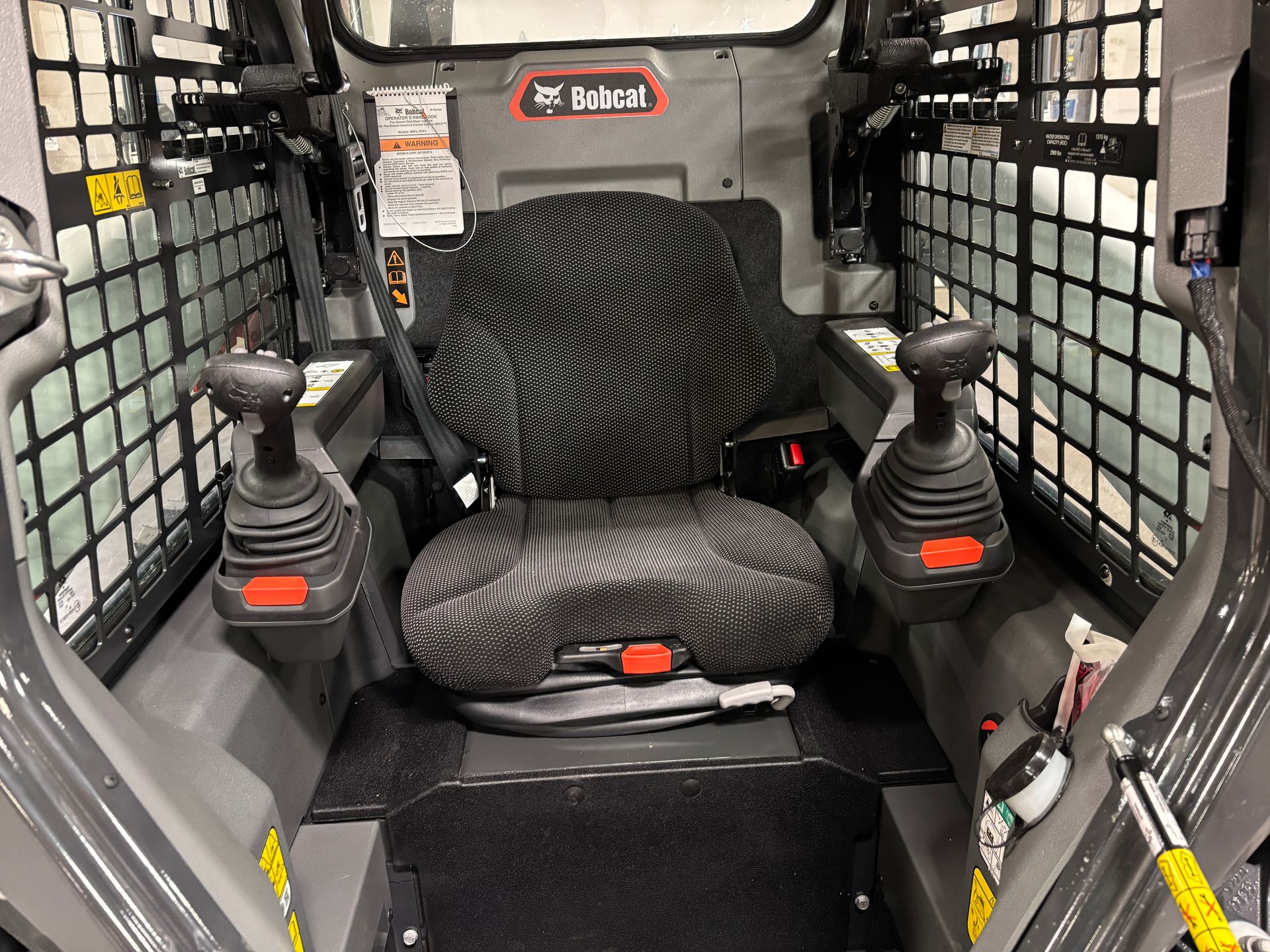Interior of a Bobcat skid-steer loader showing the operator's seat, joysticks, and safety bars.