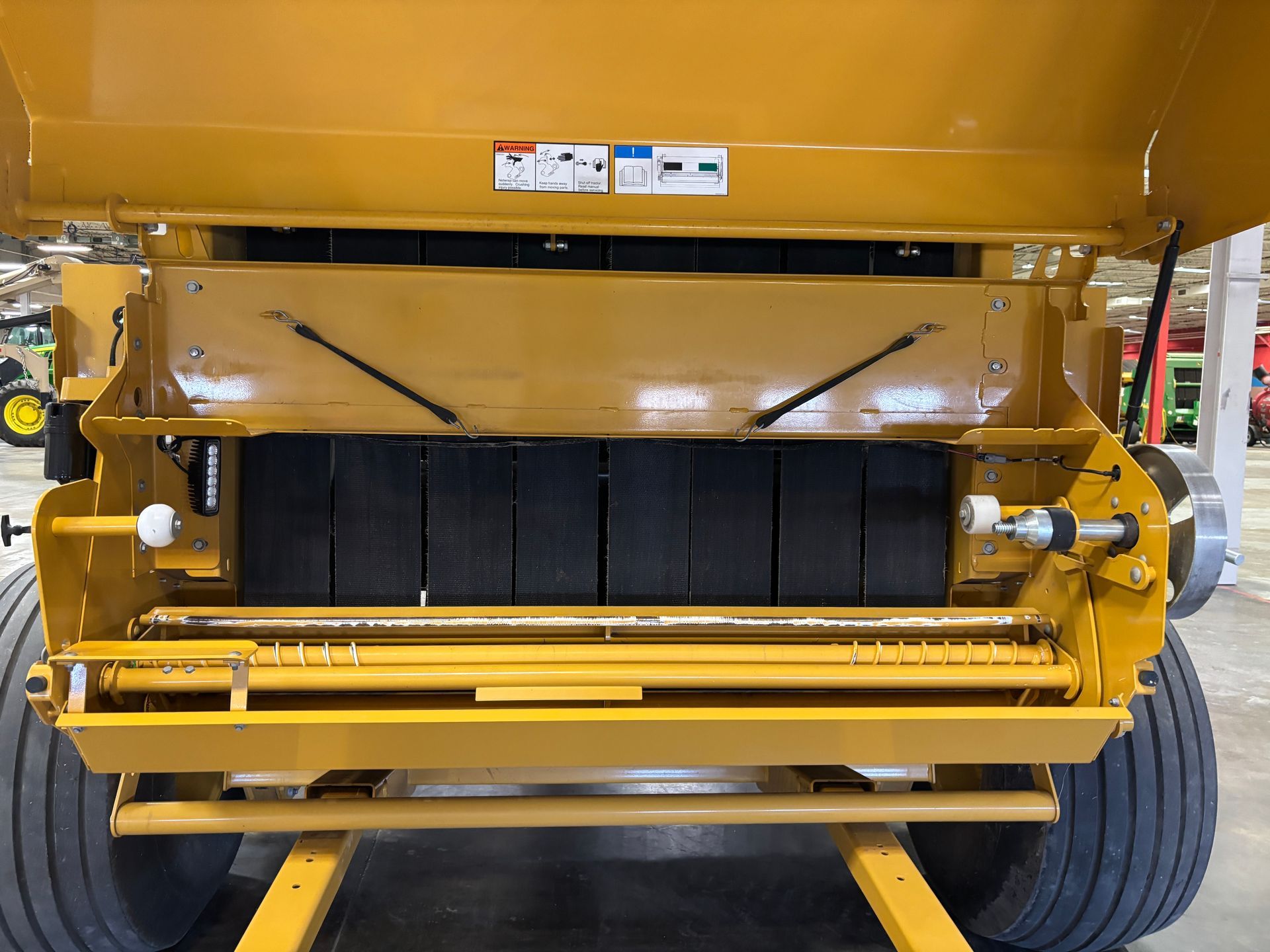 Yellow agricultural hay baler, front view; black weighted blocks, wheels, and safety signs visible.