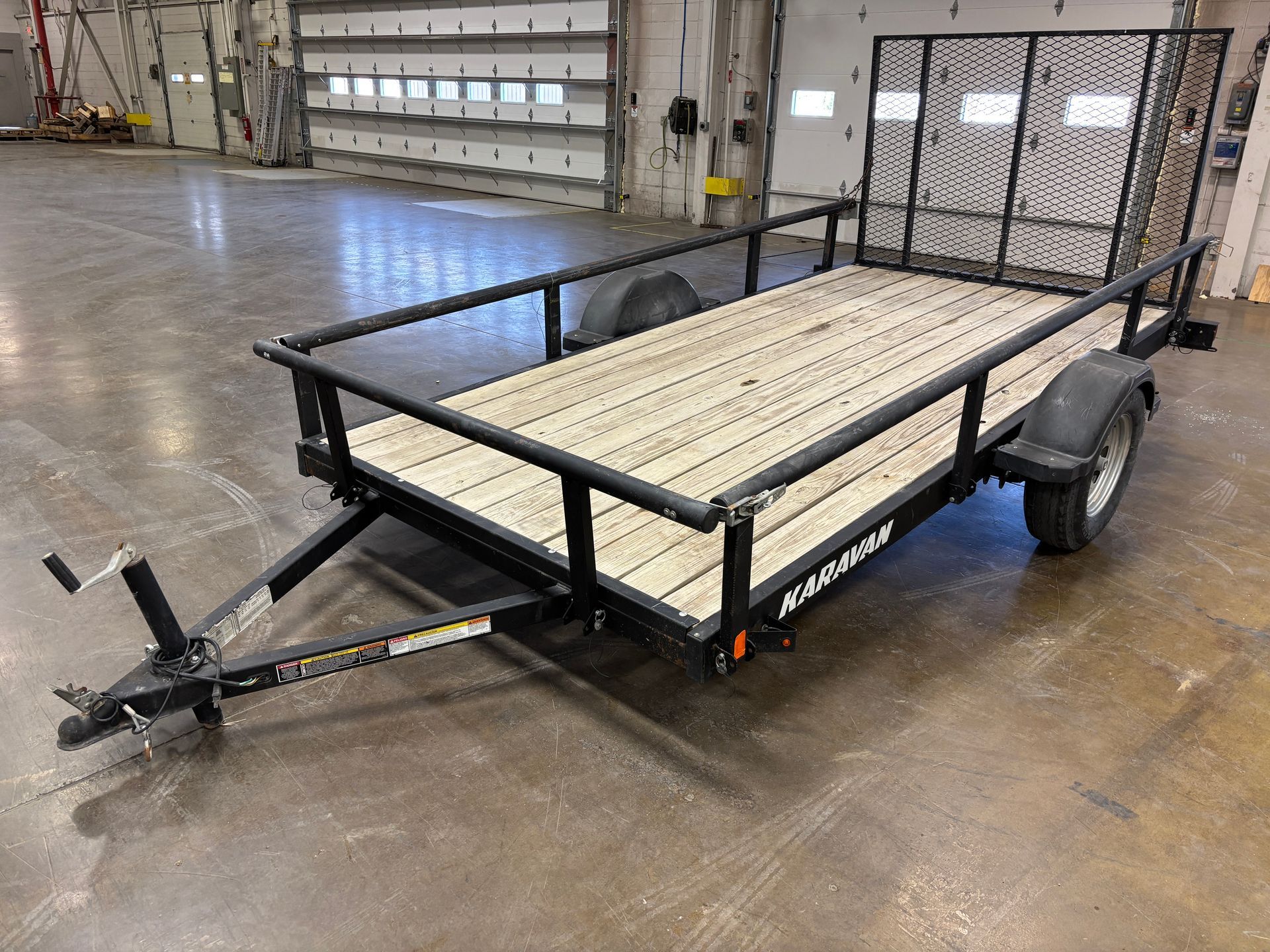 Black utility trailer with wooden deck, black mesh side and rear, inside a warehouse.