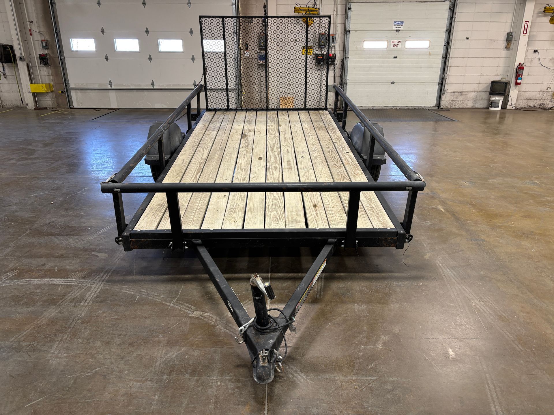 Black utility trailer with wooden deck, black railings, and metal gate, in a warehouse.