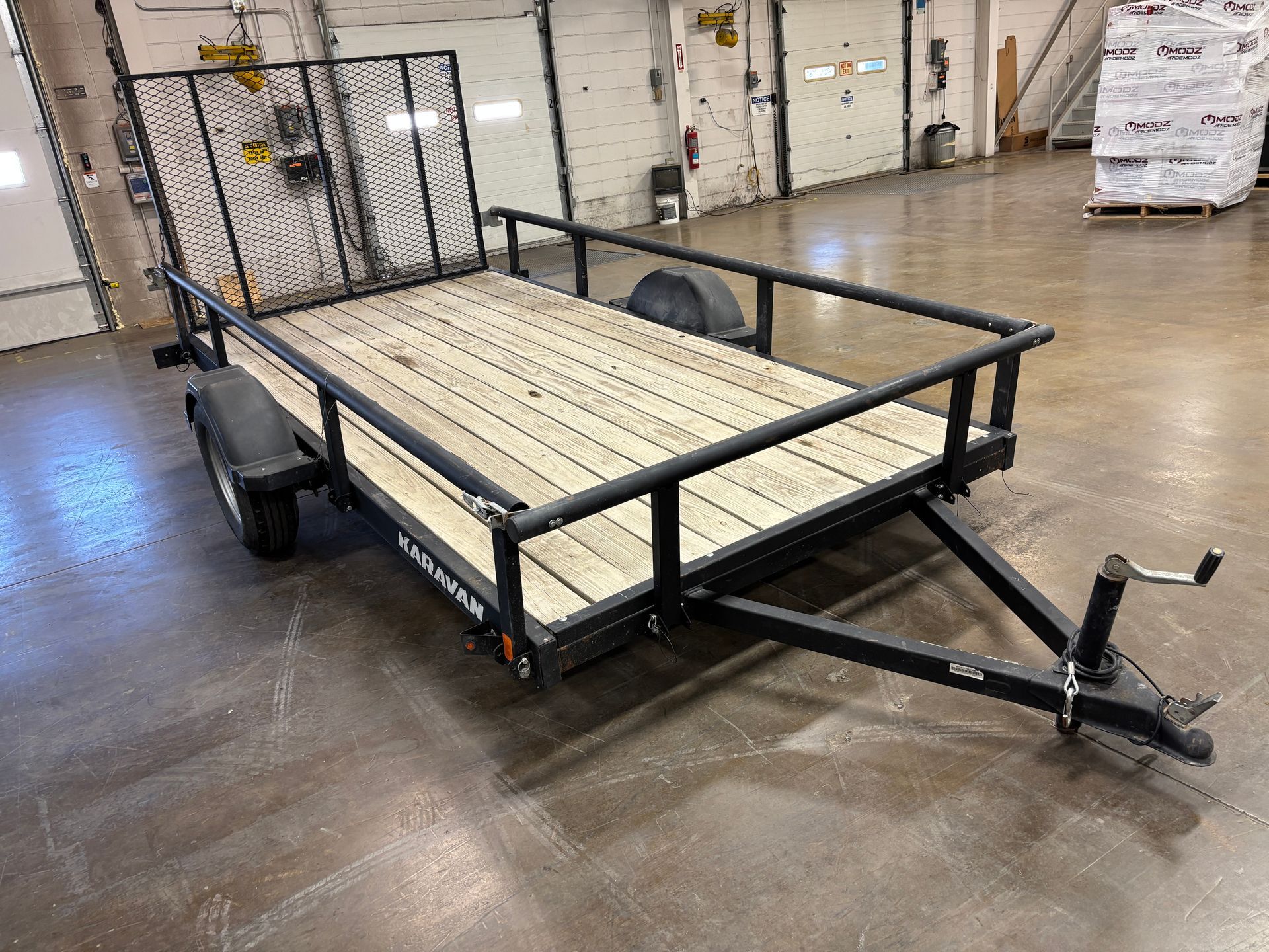 Black utility trailer with wooden deck, black frame and safety mesh, parked indoors.