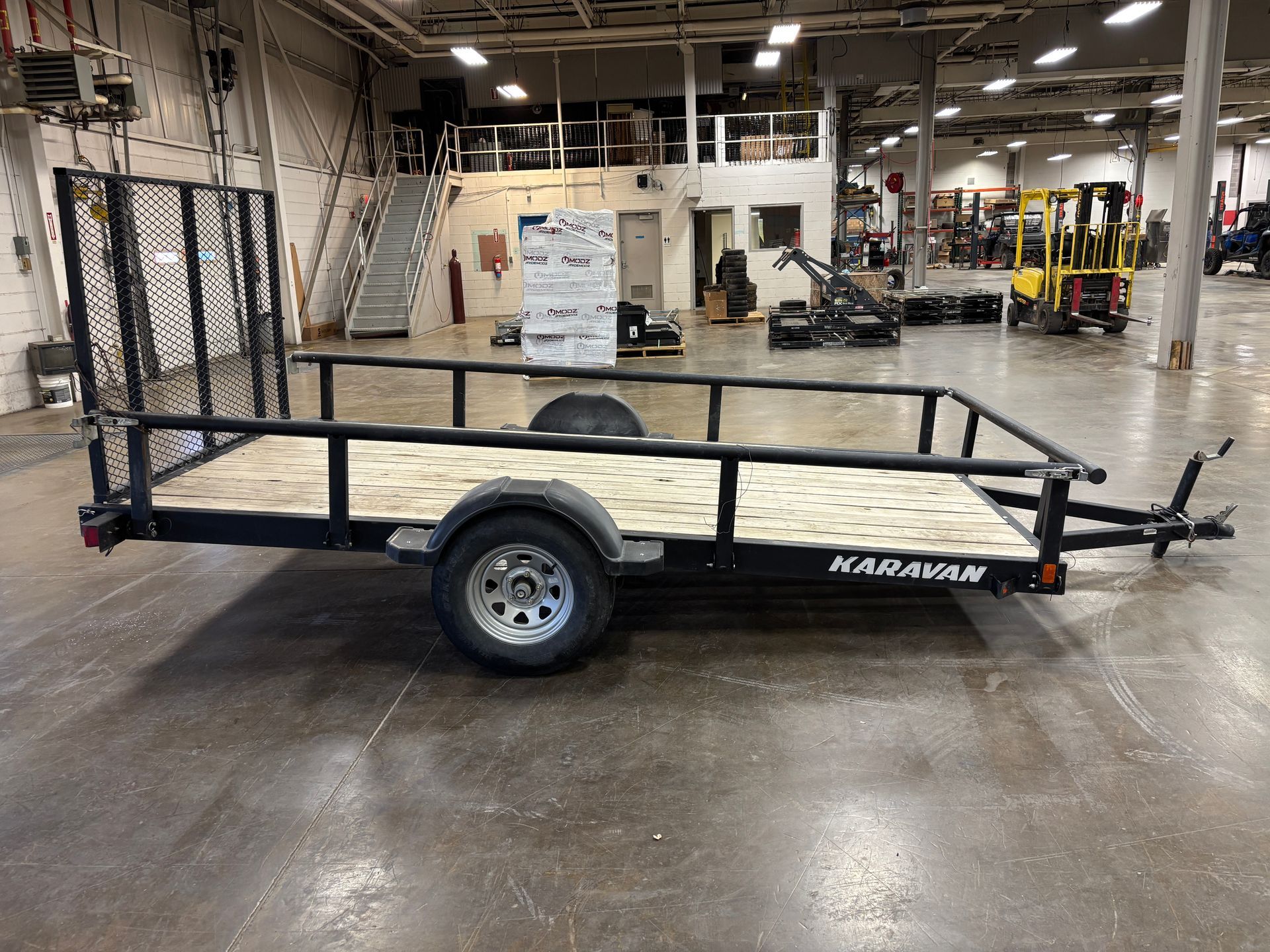 Black utility trailer with wood floor, metal side rails, and a mesh gate in a warehouse.