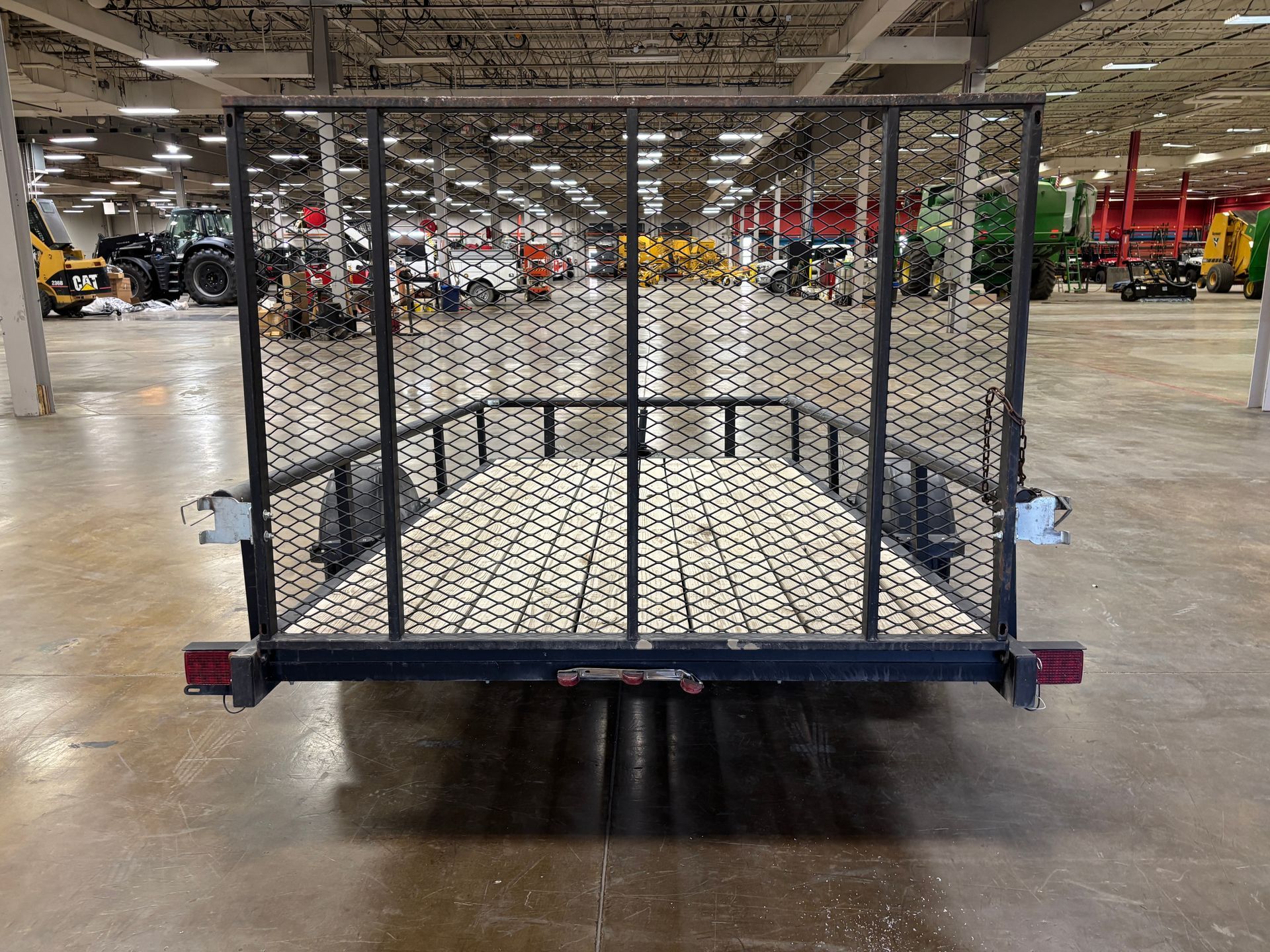 Black utility trailer with wire mesh sides, lights, and a wooden floor, inside a large warehouse.