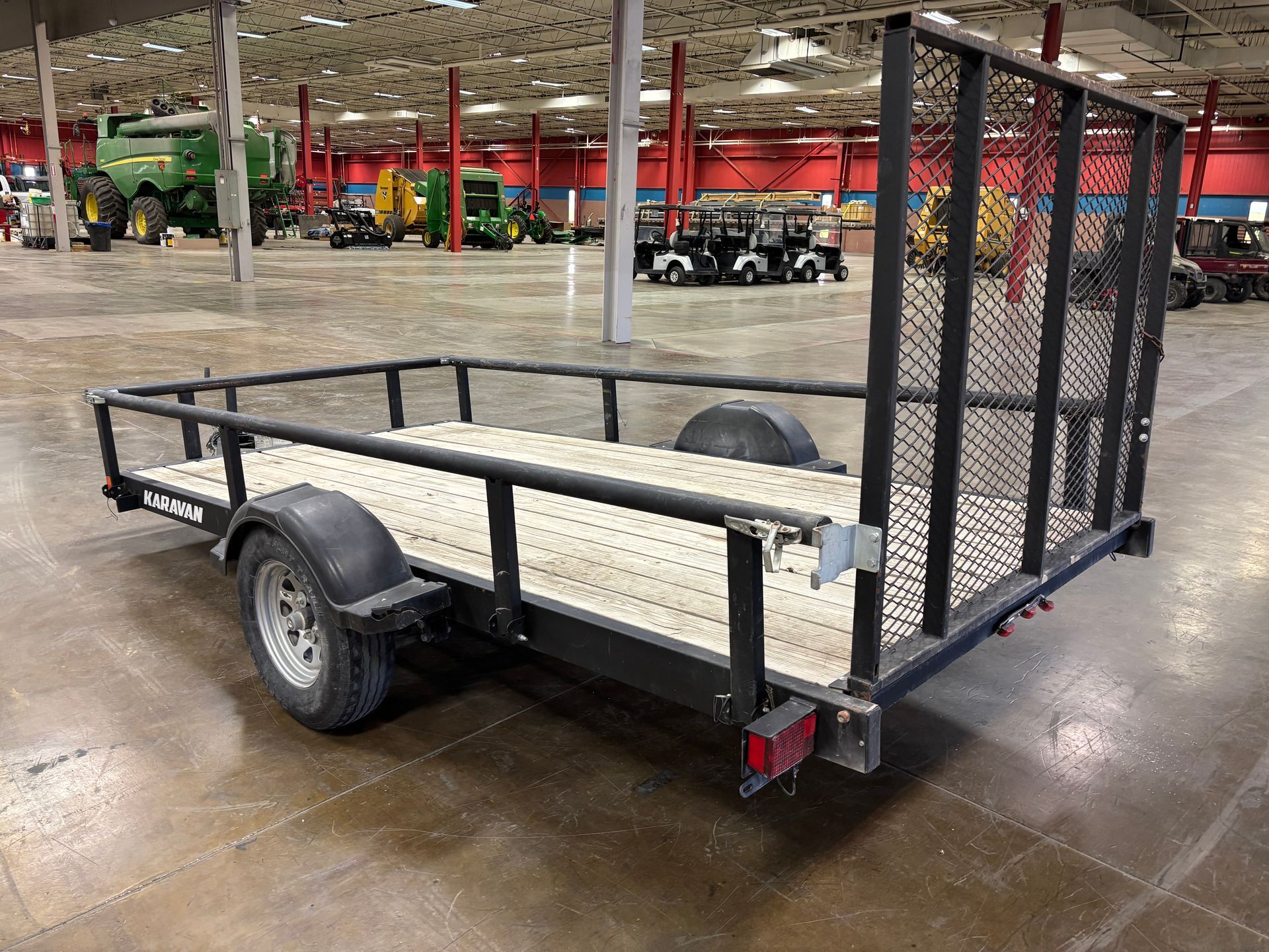 Black utility trailer with wooden bed, metal sides and mesh rear panel, inside a large building.
