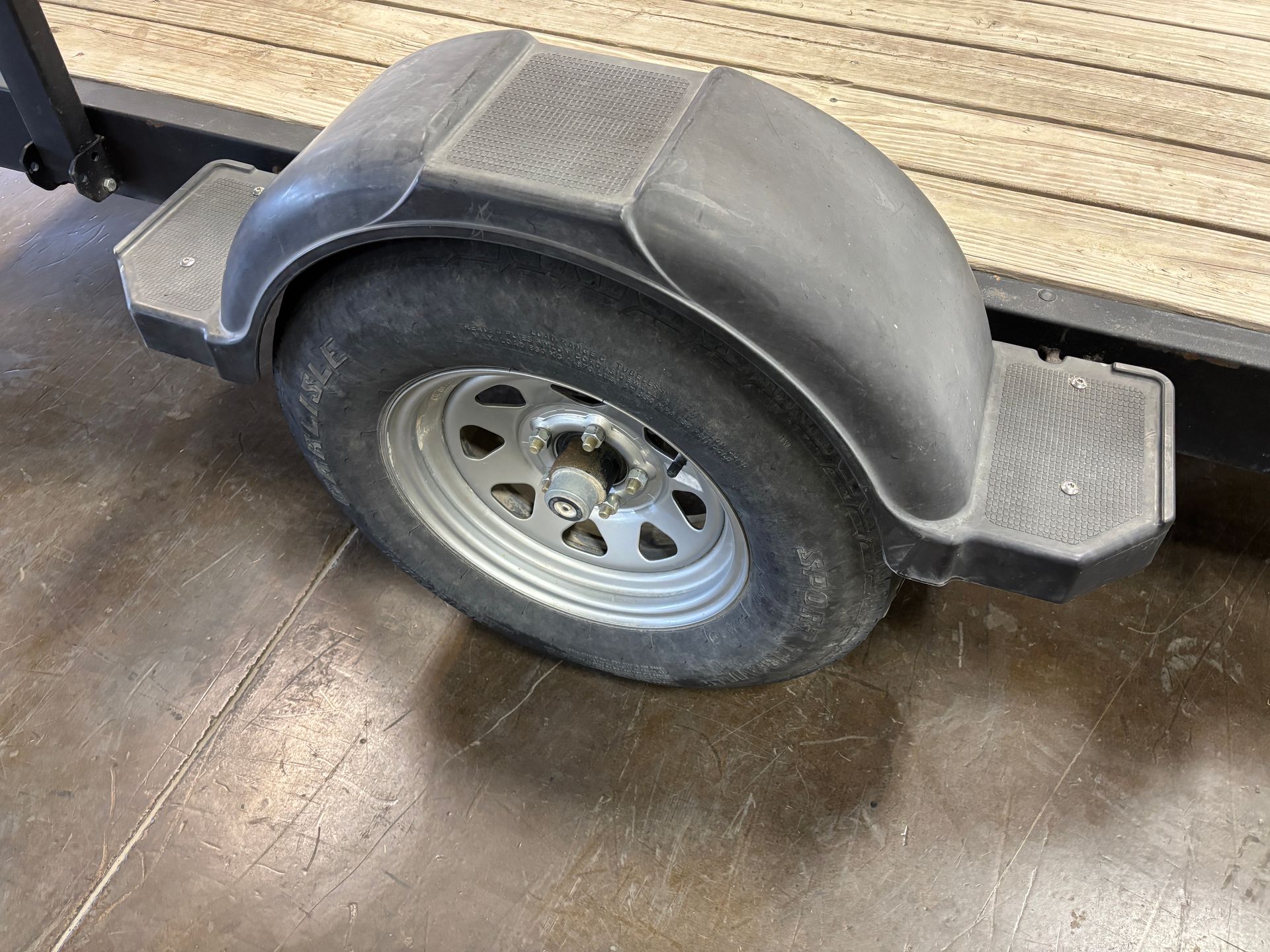 Trailer tire with black fender and silver wheel, on a wooden platform, parked on concrete.