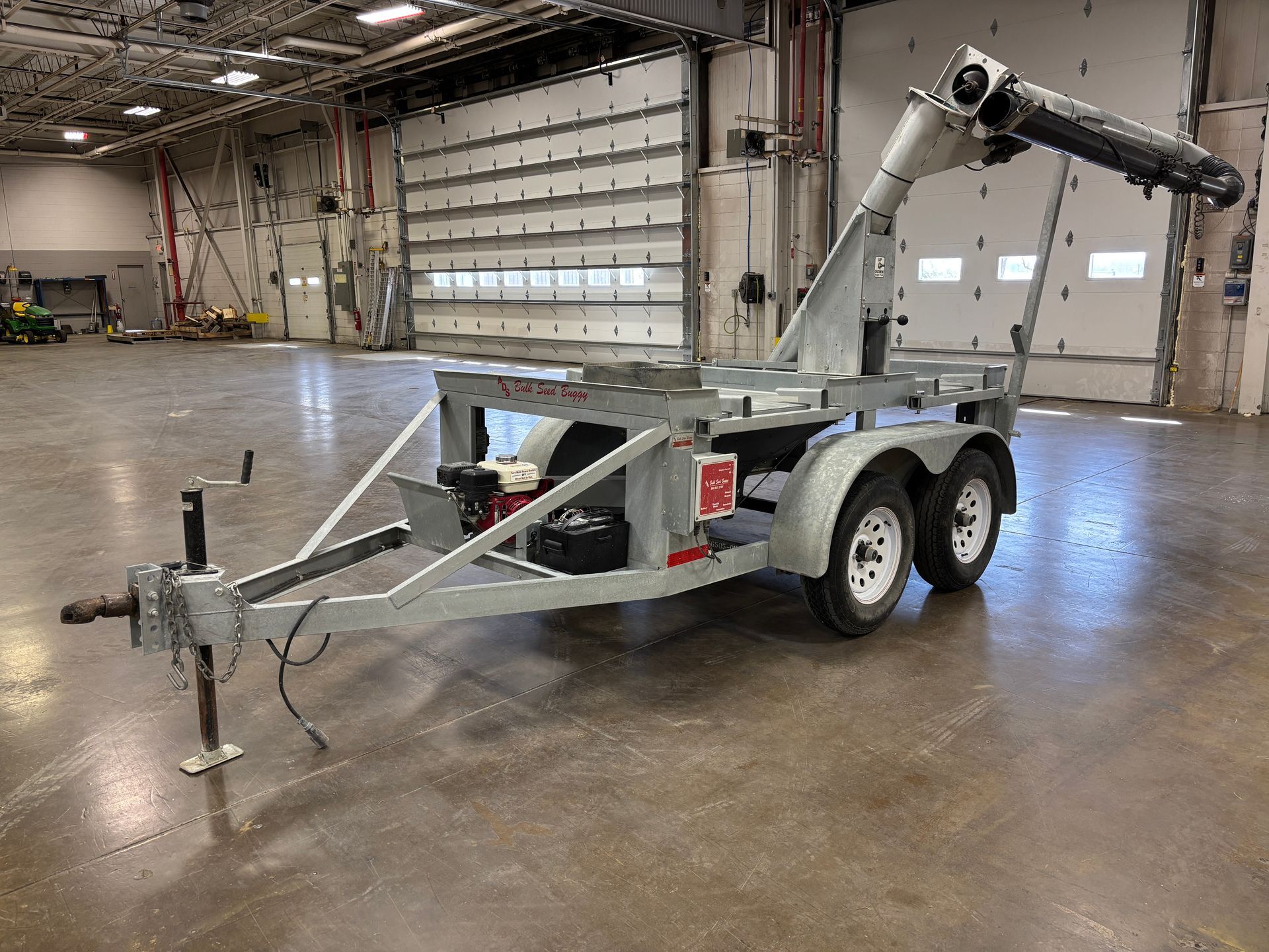 Gray utility trailer with two wheels, attached to a crane with a black arm, in a warehouse.