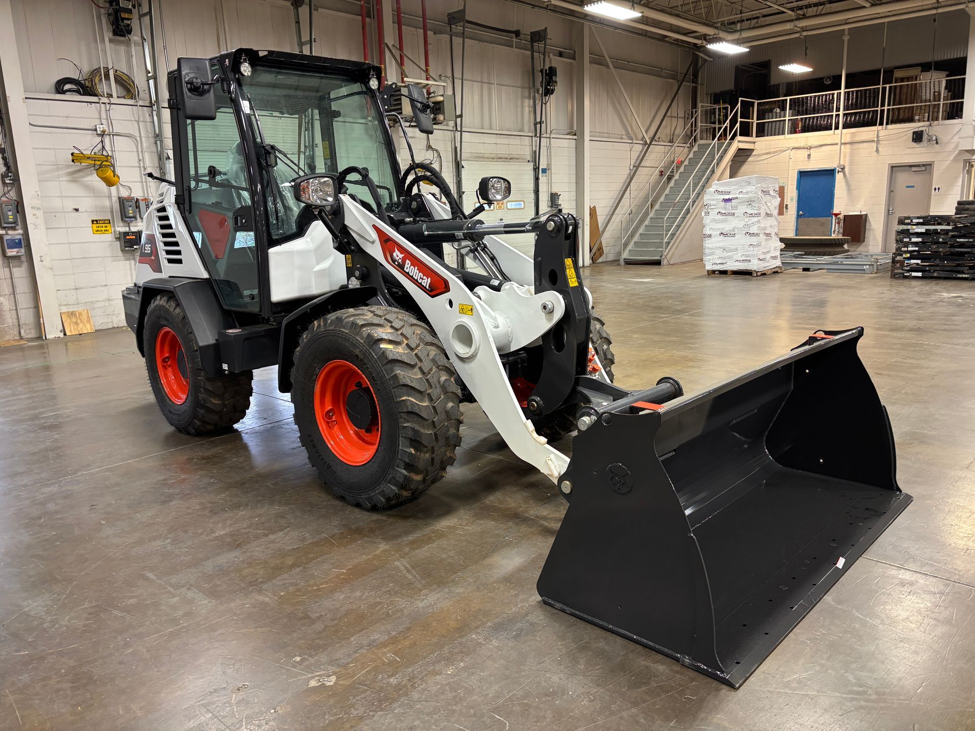 Bobcat compact wheel loader with bucket in a warehouse. White body, orange wheels, gray bucket.