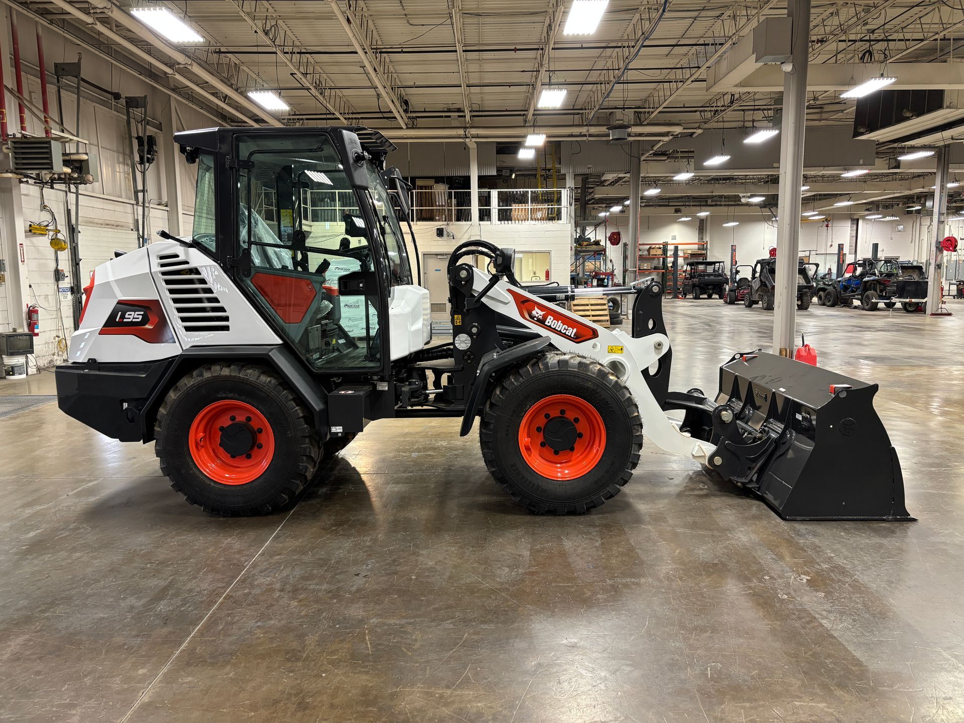 Bobcat compact loader in a large indoor space. White and black machine, orange wheels, bucket attached.